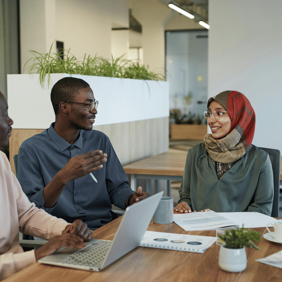 Three professionals sit around a modern office table engaged in discussion, with a laptop, papers, and a small plant on the table, surrounded by glass walls and office furniture.