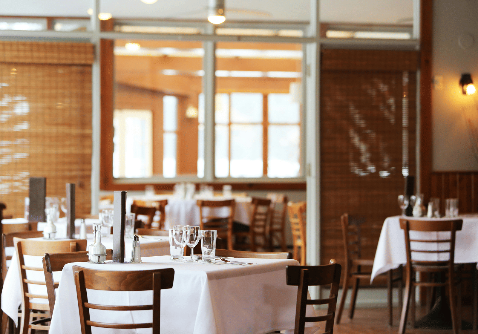 The bright interior of a restaurant with wooden tables and chairs, crisp white tablecloths, and bamboo window blinds.