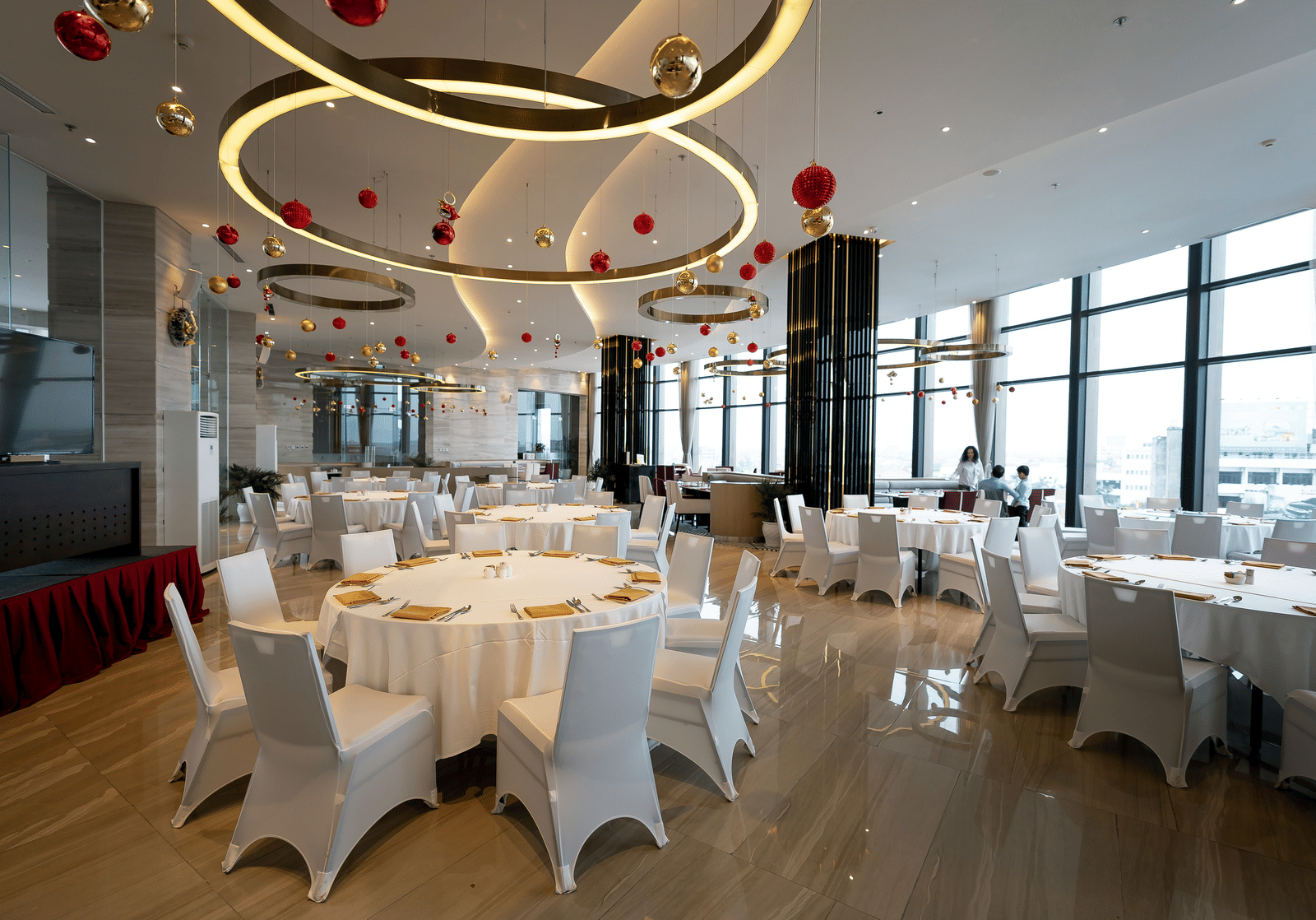 A bright, spacious event hall set up for a reception with many round tables, white chair covers, and modern circular ceiling lights.