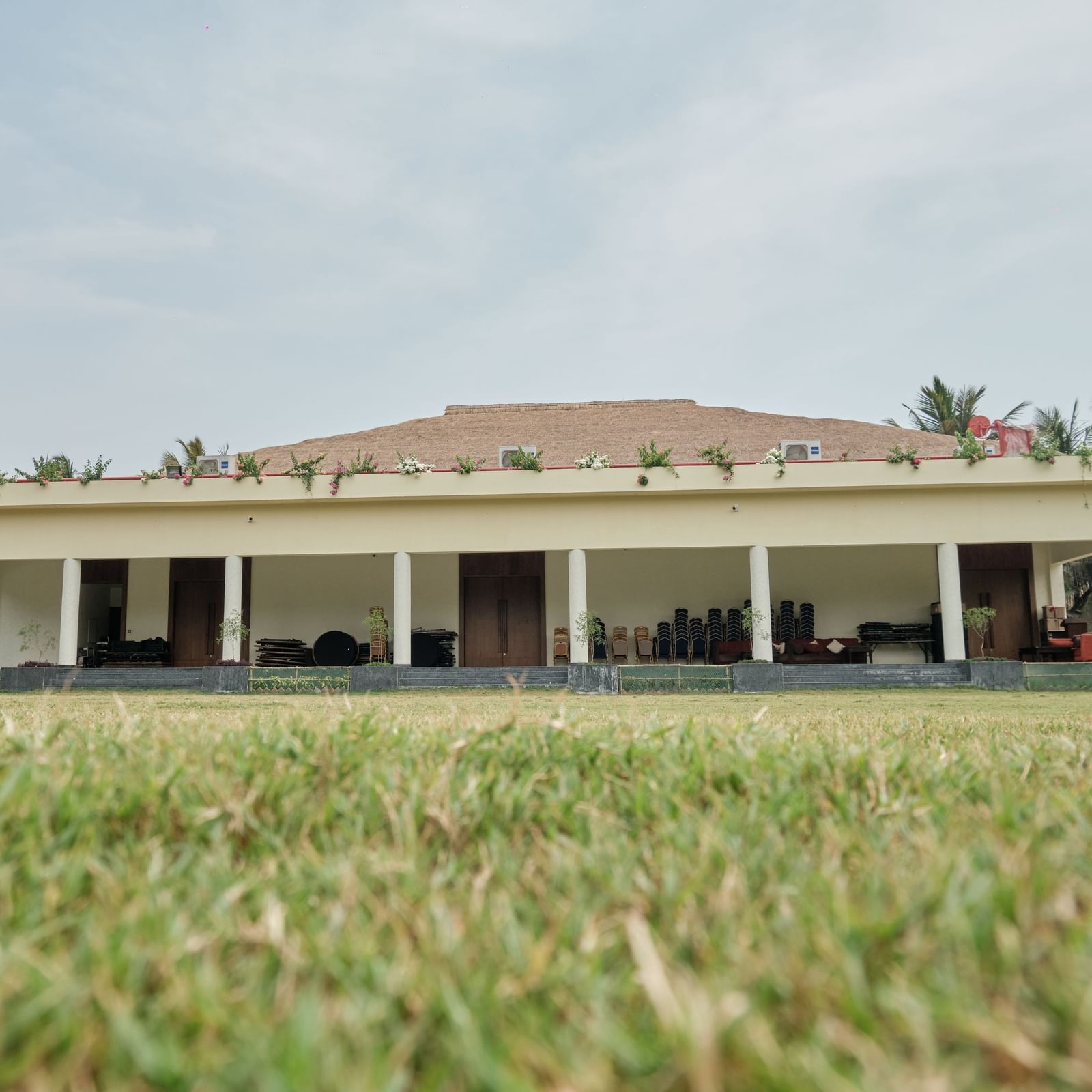 A long, single-story building with a covered veranda stretches across a green field under a clear sky | Utsav lawn