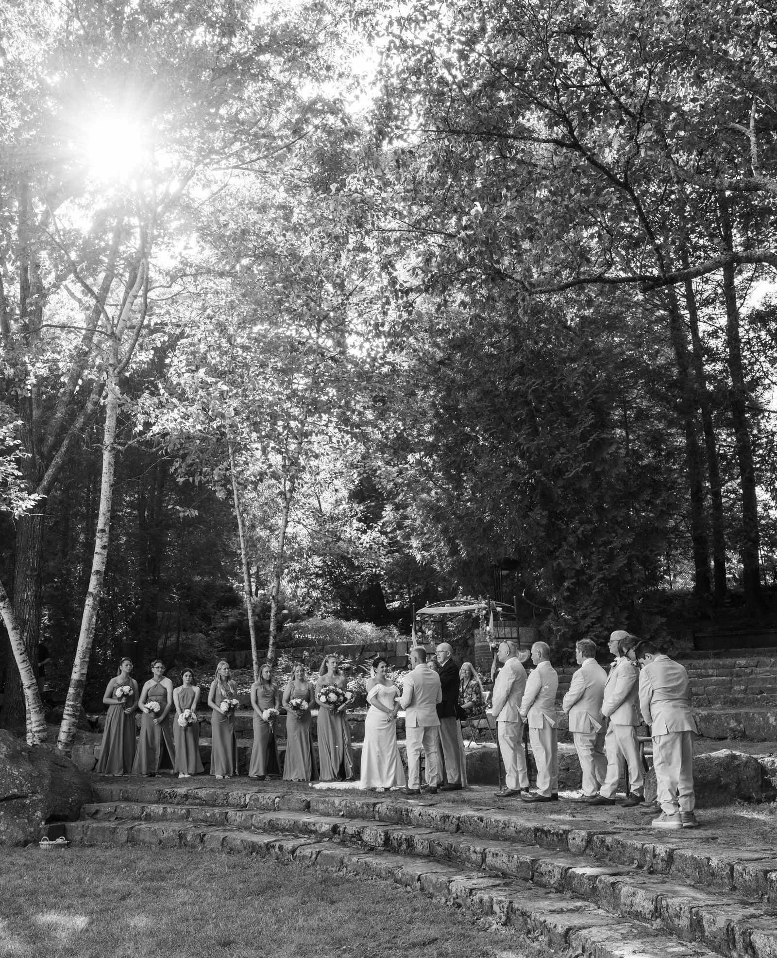 Wedding ceremony at Hartstone Inn with the couple, officiant, bridesmaids, and groomsmen standing on stone steps surrounded by trees and sunlight filtering through.