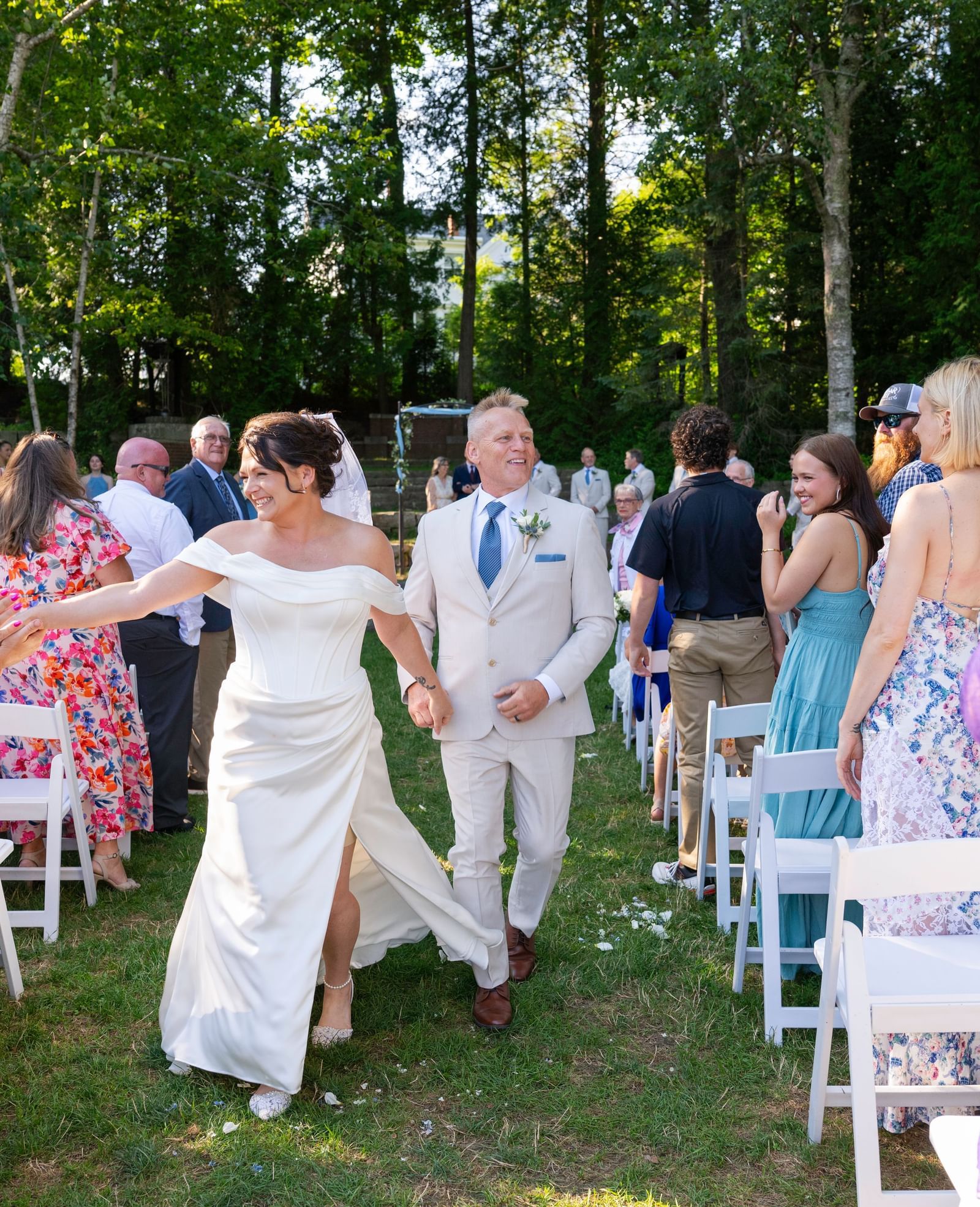 At Hartstone Inn, a newlywed couple walks down the aisle holding hands, surrounded by standing guests who are smiling, clapping, and reaching out to greet them.