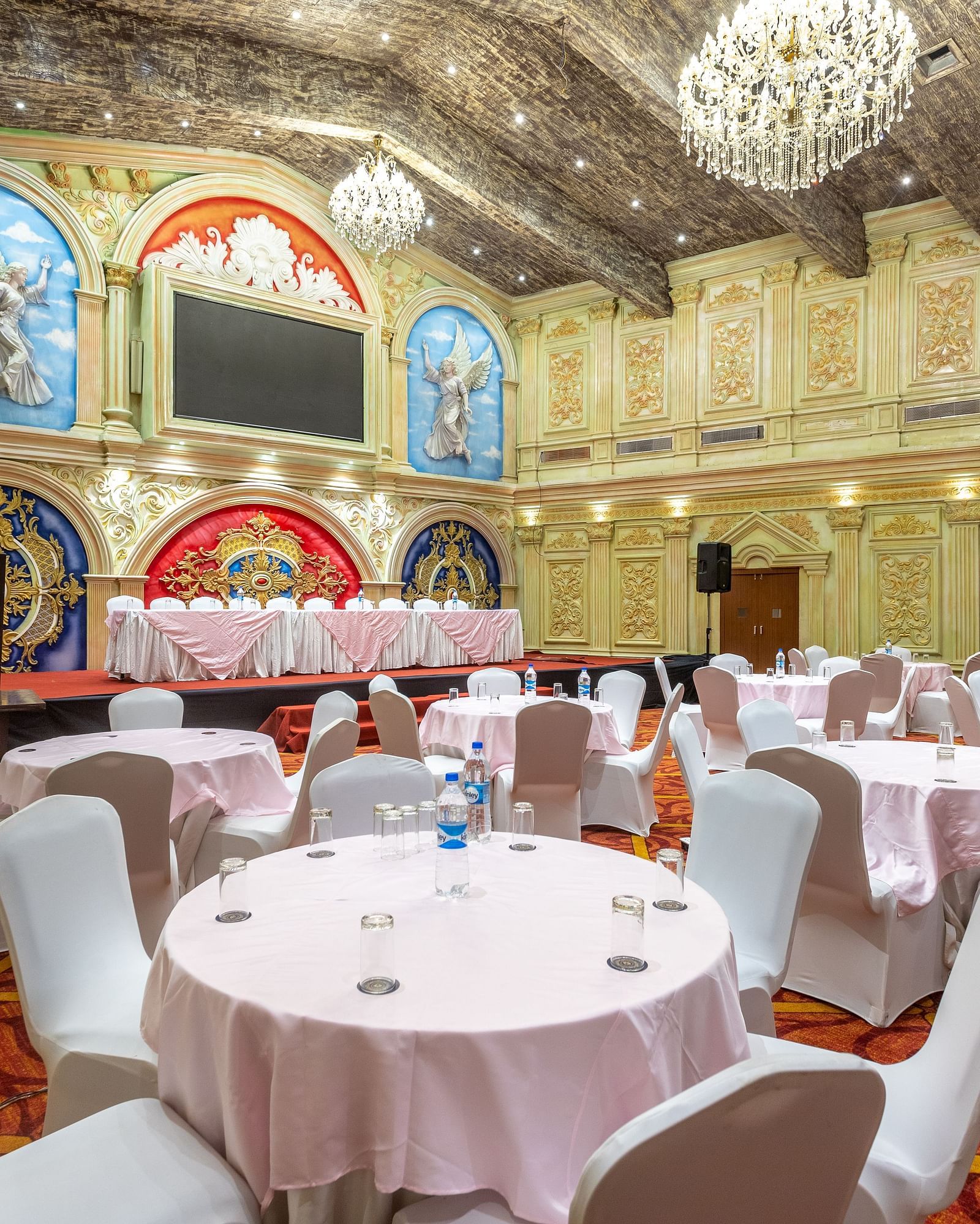 Ornate banquet hall at Diamond Palace Lords Plaza with round tables, chairs, chandeliers, and decorative walls.