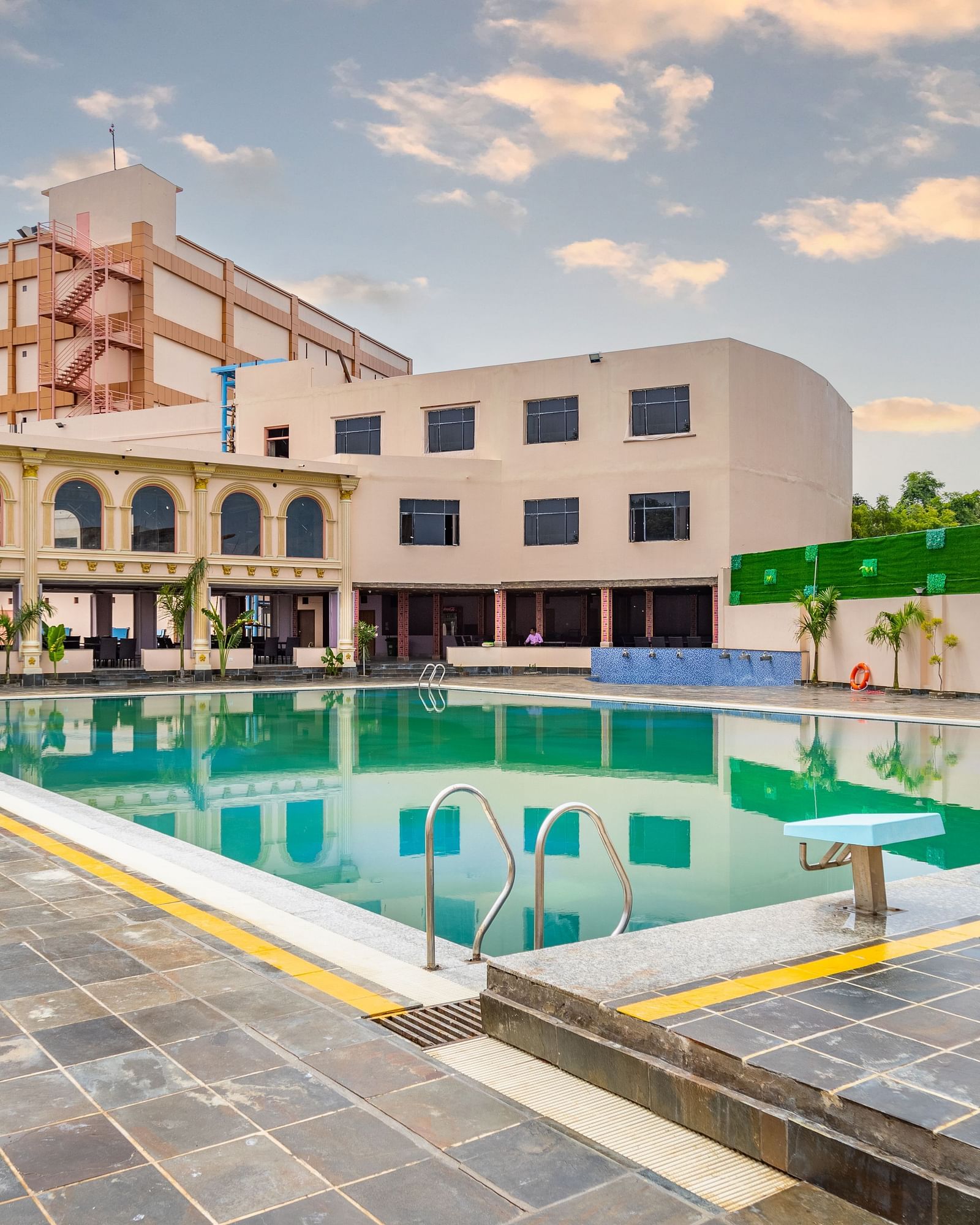 Hotel pool reflects buildings under a cloudy sky with in-water seating and tiled surroundings.