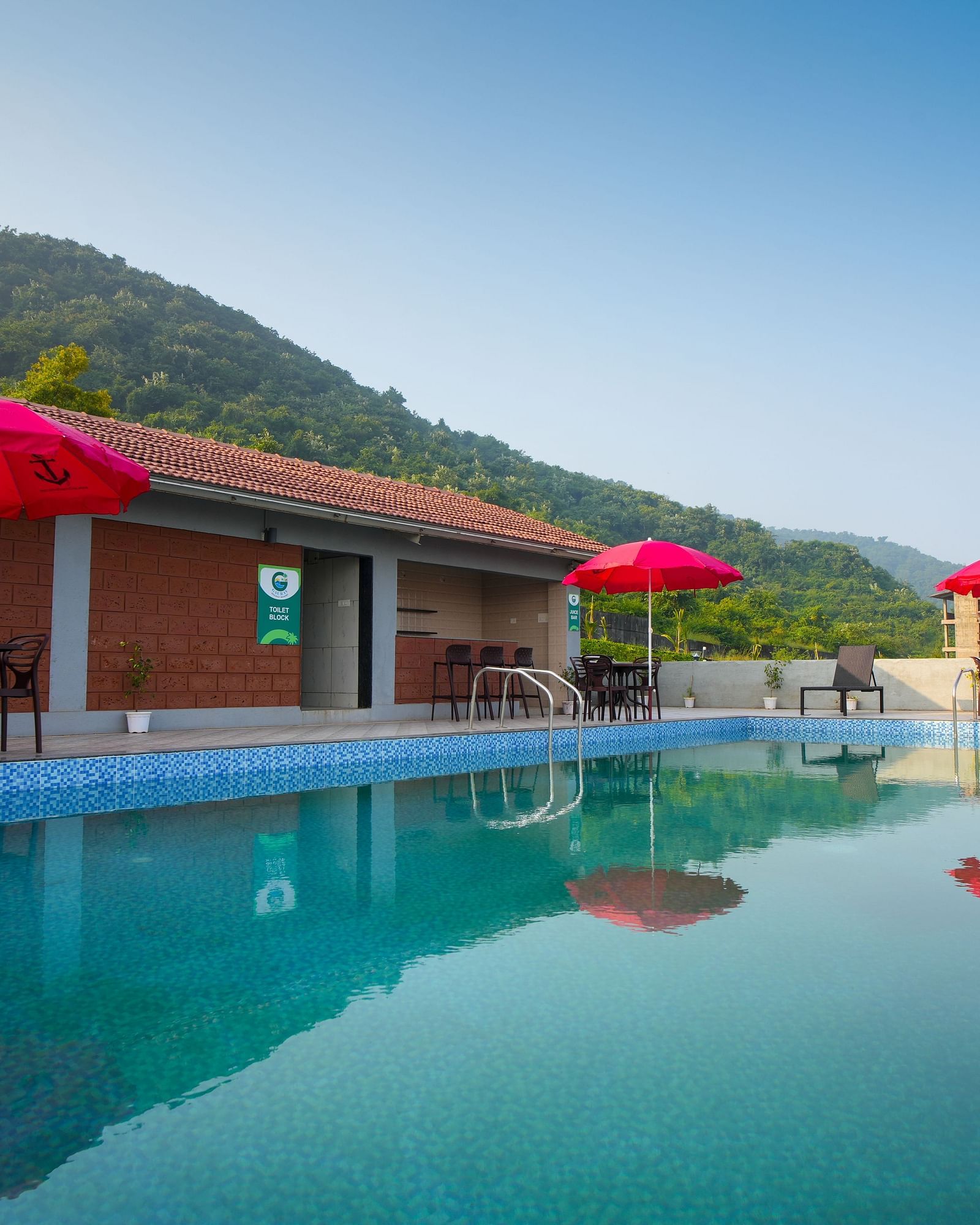 A swimming pool with red umbrellas and lounge chairs, set against a backdrop of mountains and a building - Gaurav Lords Resort Shrivardhan.