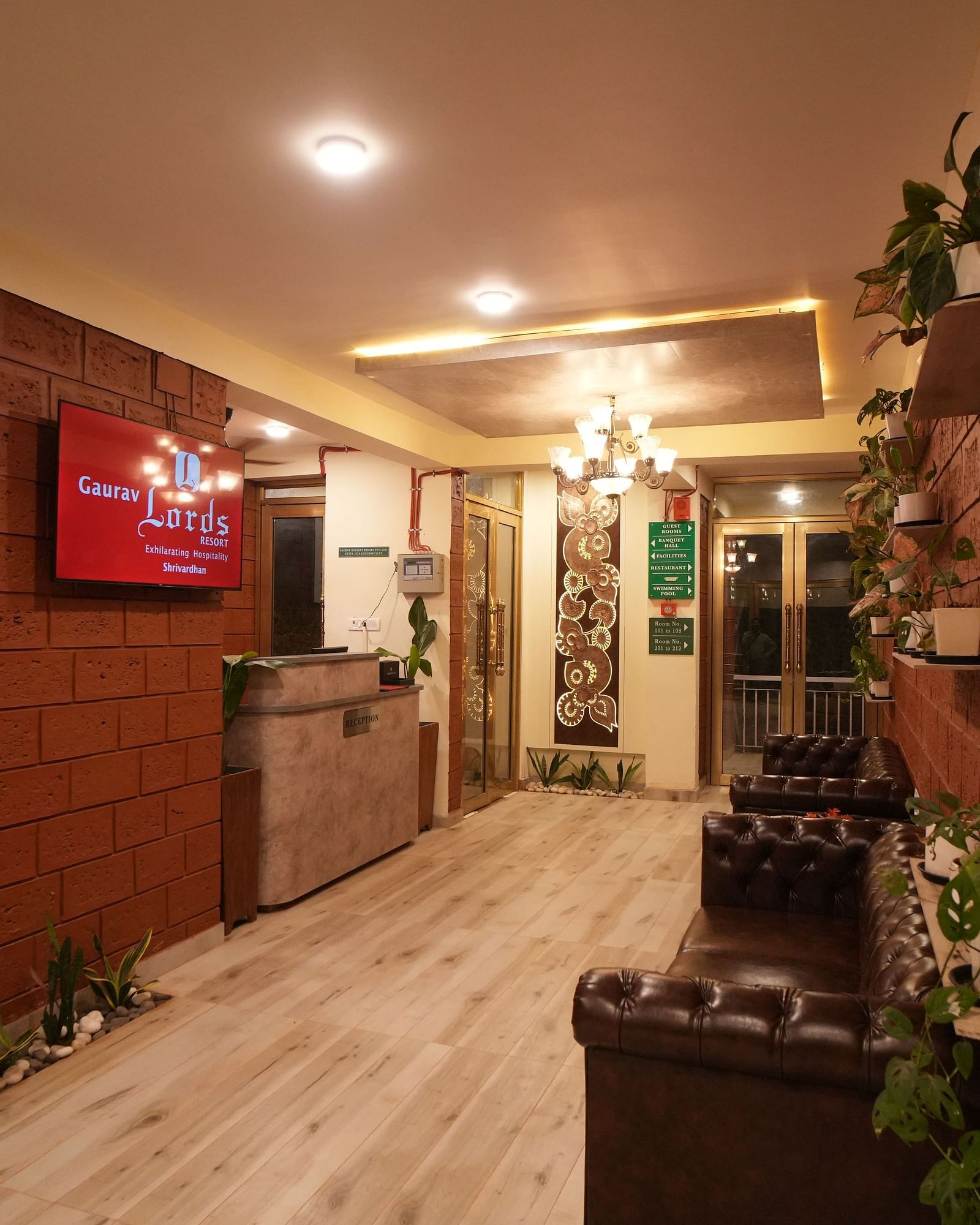 Interior hallway of a resort with textured red brick walls, a wooden reception desk, a dark leather sofa, and potted plants. A television is mounted on the wall - Gaurav Lords Resort Shrivardhan.