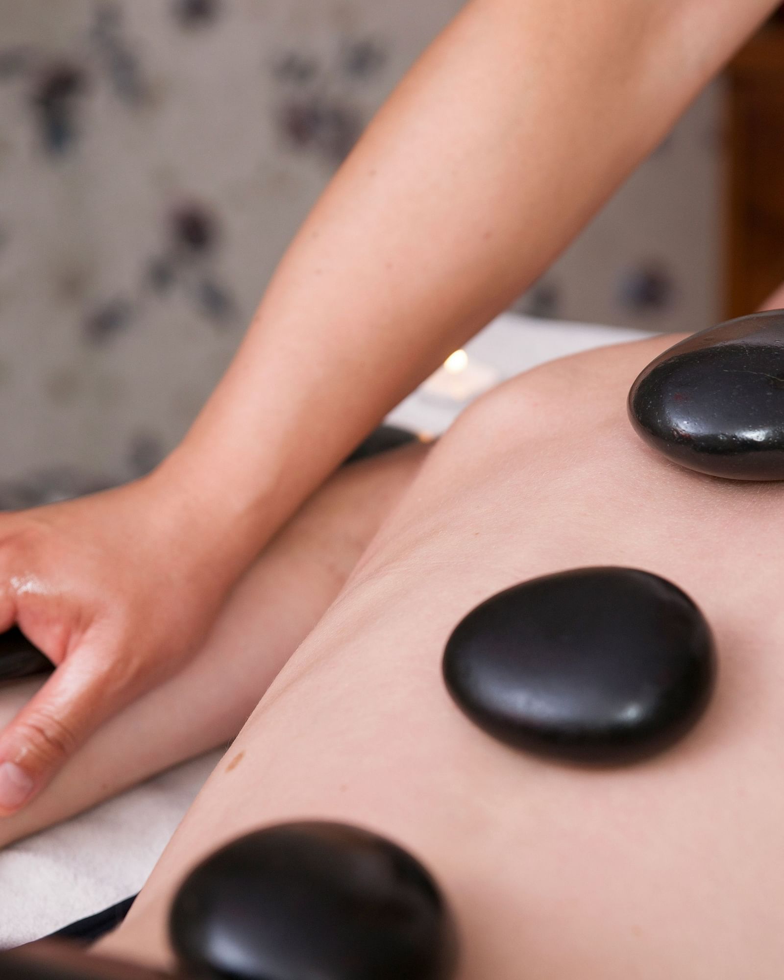 A person receiving a hot stone massage on their back, with smooth stones placed along their spine and a therapist's hands working on their shoulder. A flower adorns the person's hair.