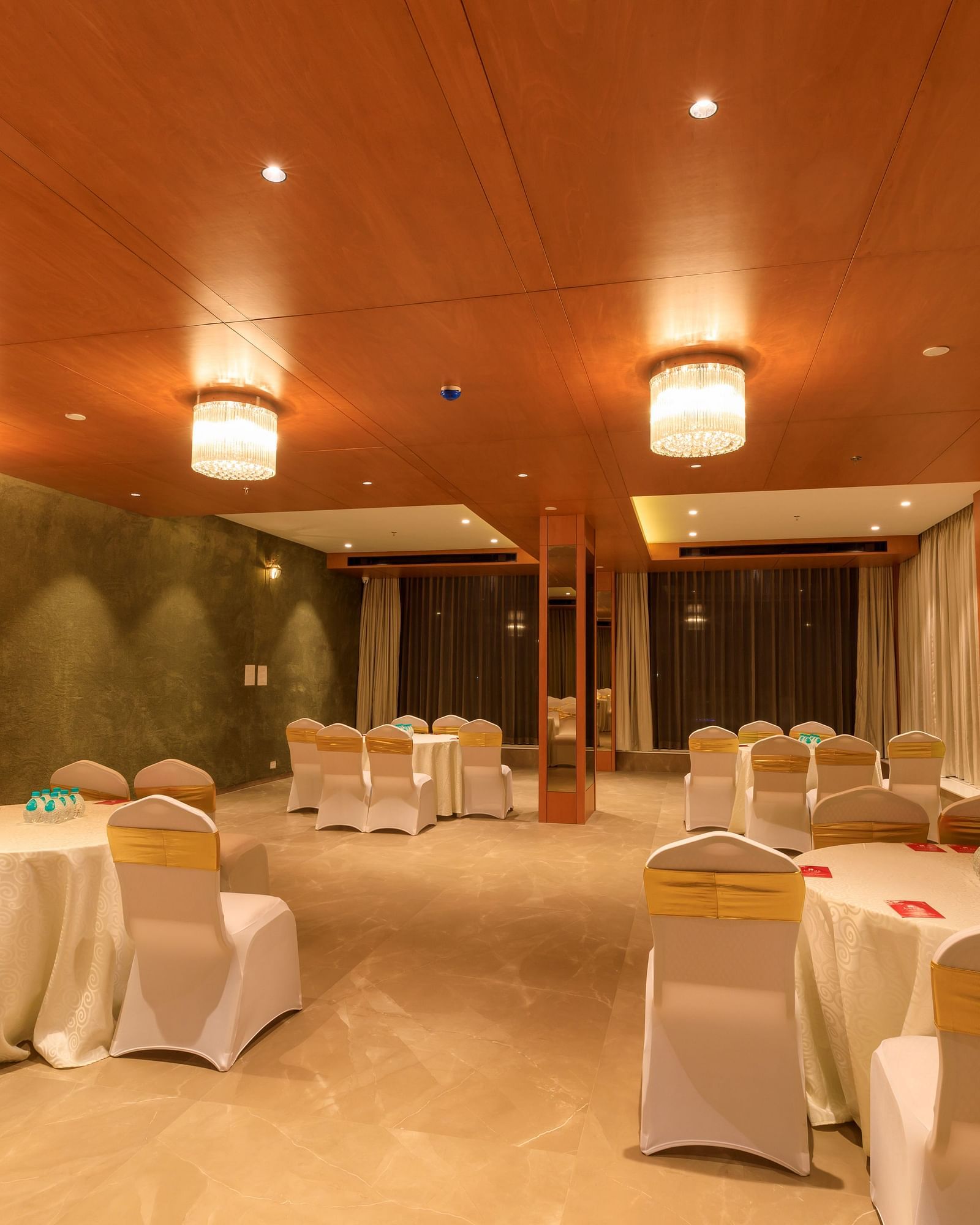 A warmly lit banquet hall at Lords Eco Inn Apple Gate is set with round tables draped in white, surrounded by chairs with gold sashes.