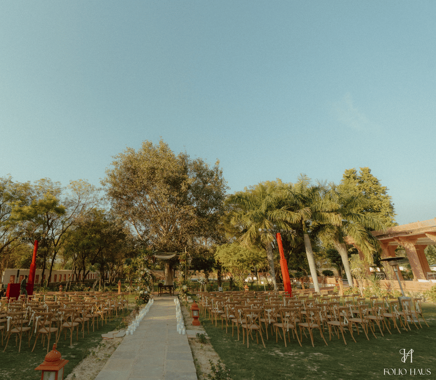 An outdoor wedding ceremony setup at Umaid Palace with wooden chairs arranged on either side of a central paved aisle on a spacious green lawn surrounded by trees.