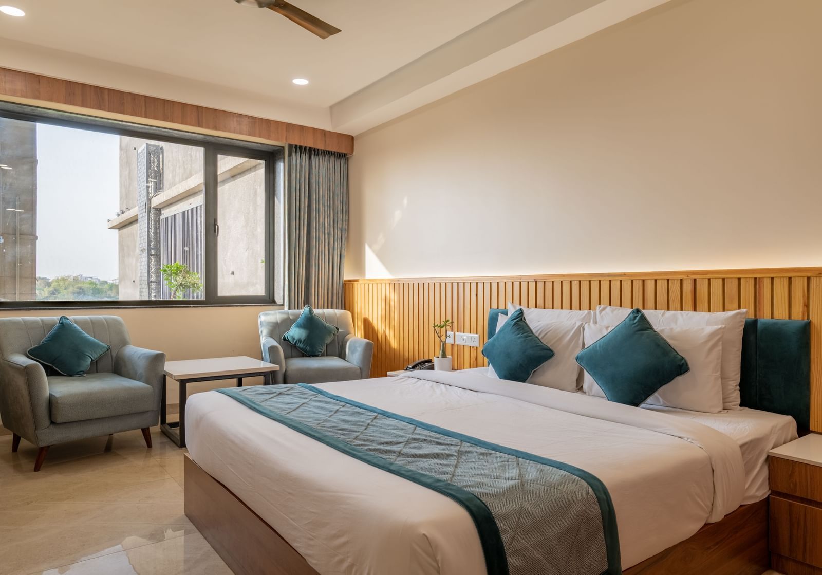 A bedroom at Hotel Highway King, Bagru, with bedside tables, armchairs, large window, and decorative cushions on bed.