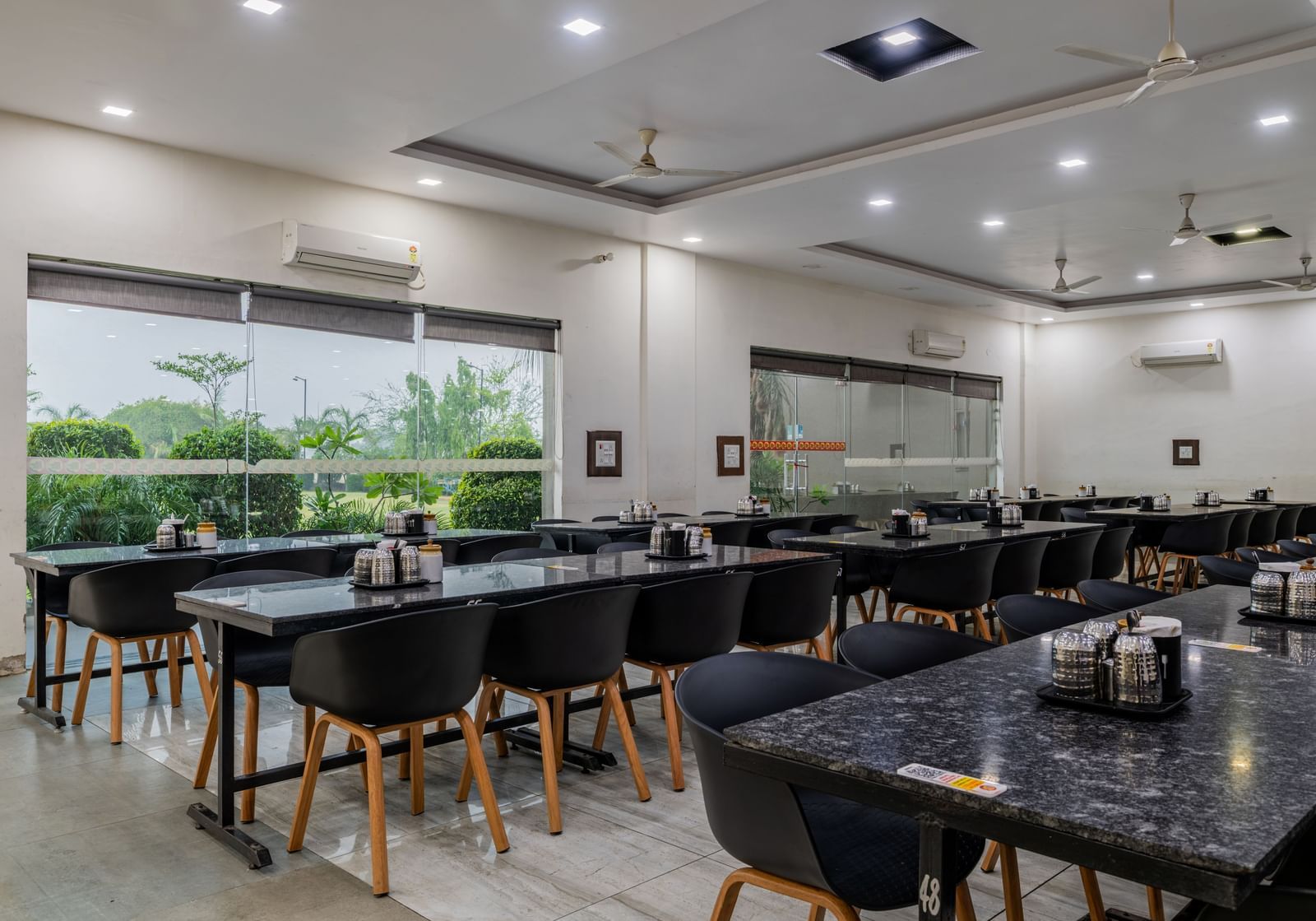 Air-conditioned restaurant of Hotel Highway King, Behror, is furnished with parallel aligned dining tables, water cups and glass windows that offer views of the trees outside at Hotel Highway King, Behror.