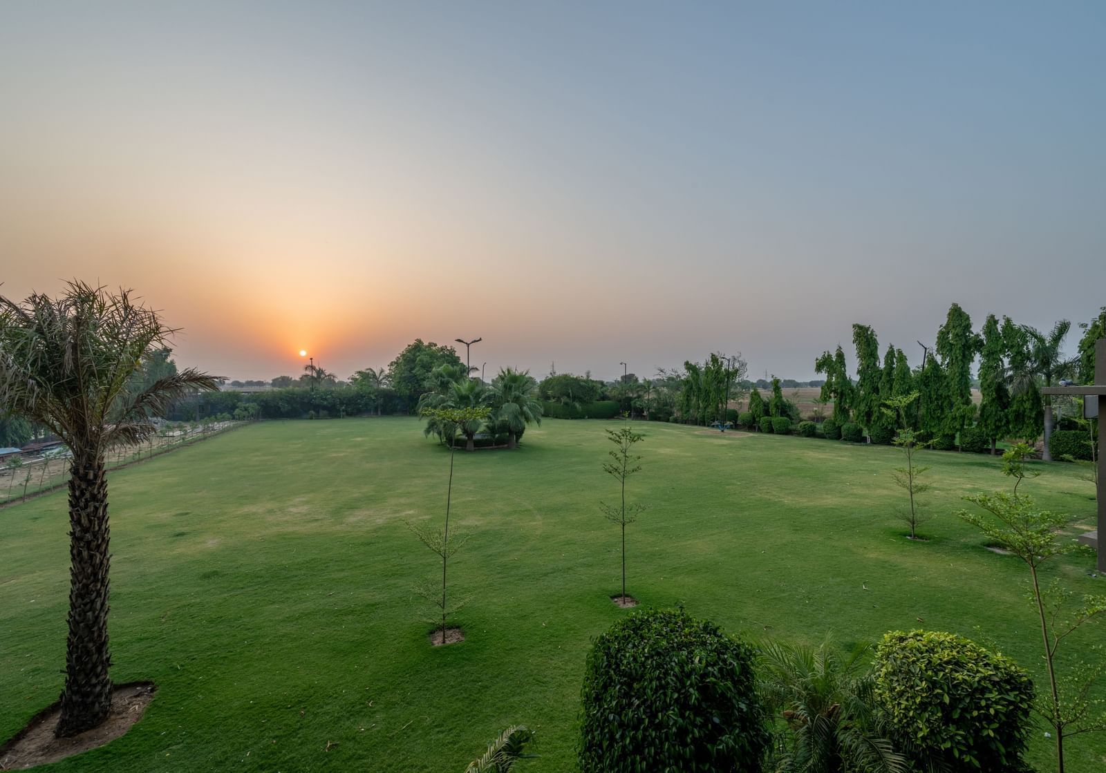 An extended view of the expansive open garden area in the premises of Hotel Highway King, Behror, seen with trees, green grass, and glowing sun in the background.