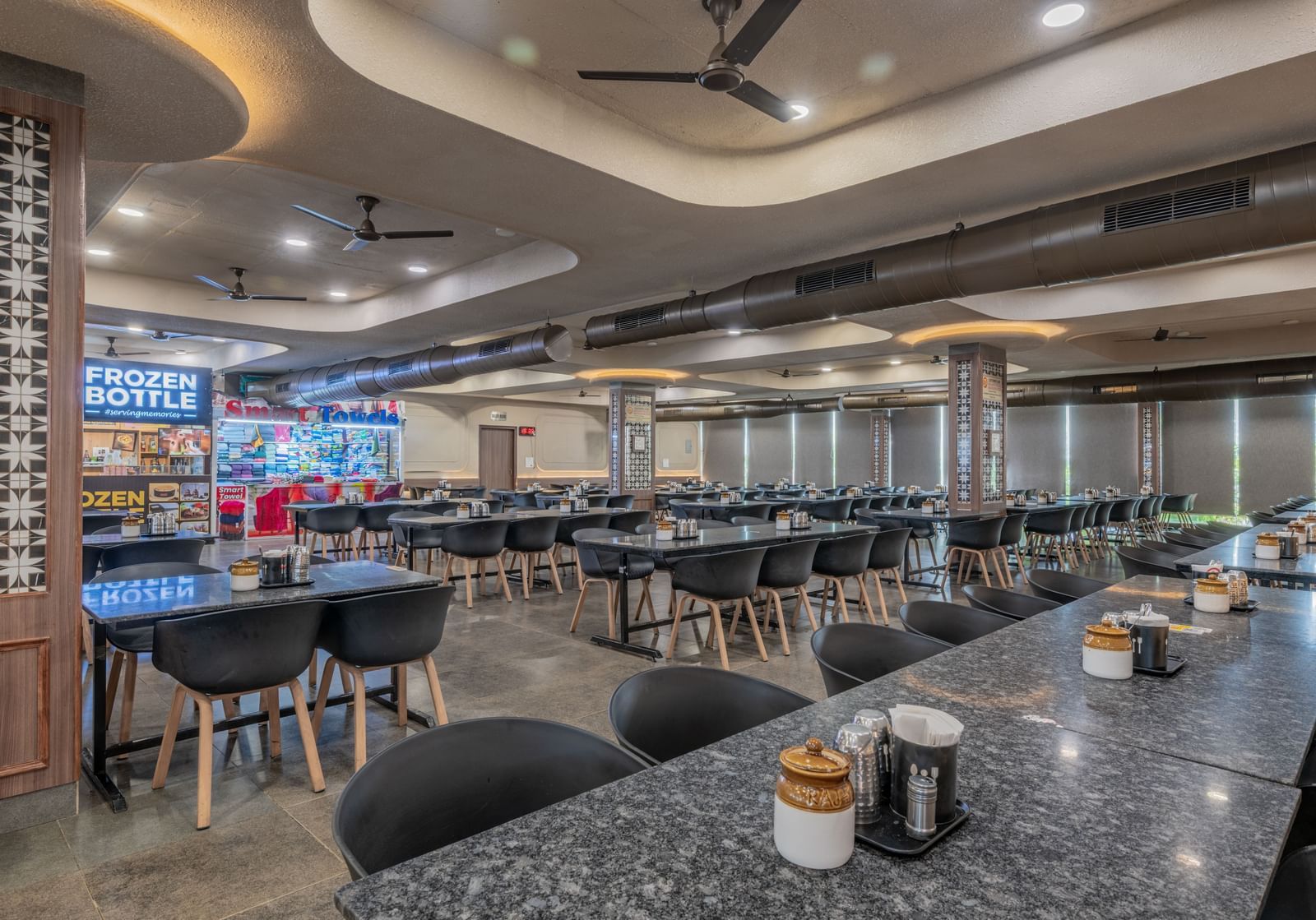 A restaurant interior with sleek tables, ambient ceiling lights, and neatly arranged seating at Hotel Highway King, Neelka.