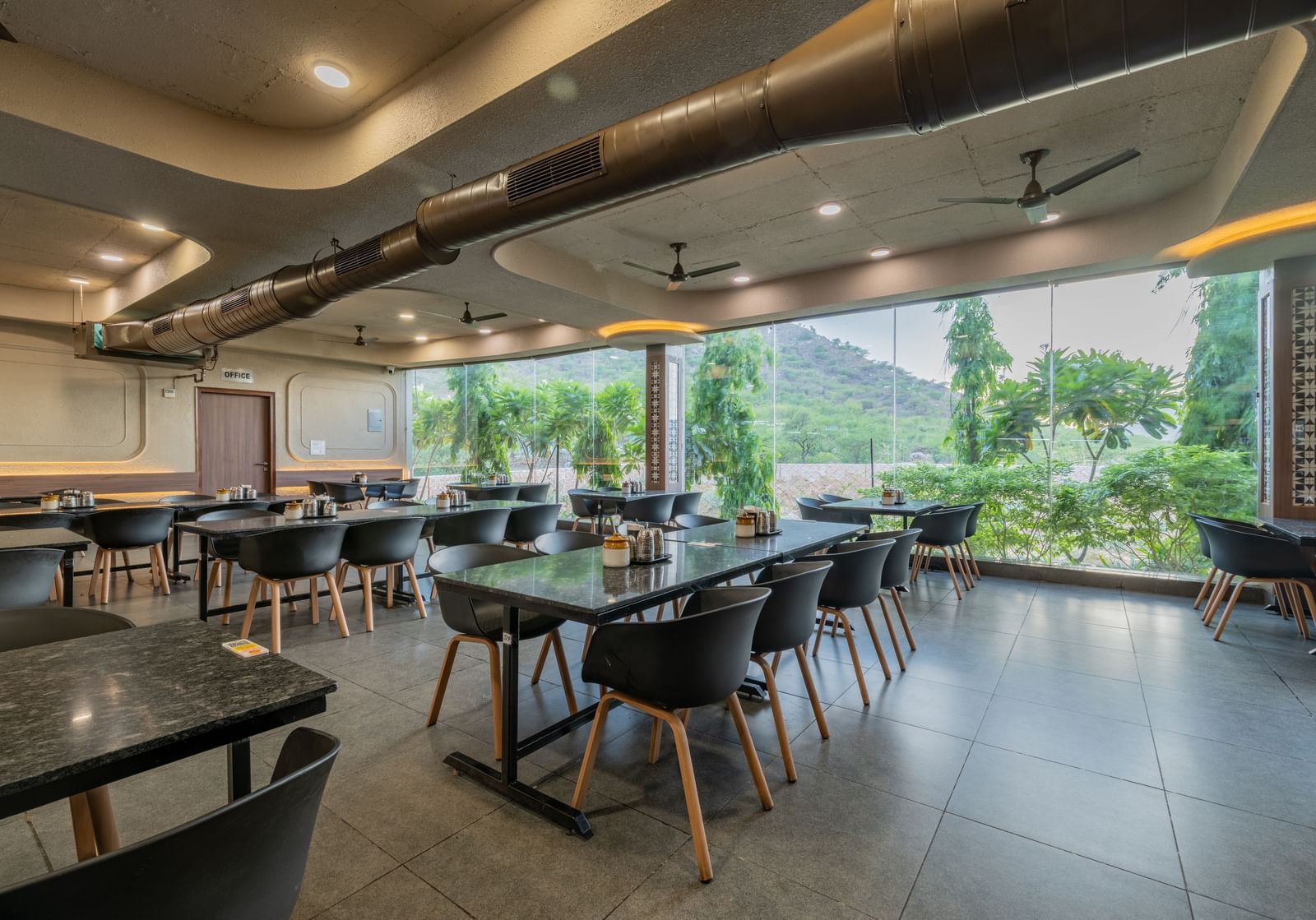 Diagonal view of the restaurant interior at Hotel Highway King, Neelka, with glass panels behind the seating area overlooking the lawn and lush mountains.