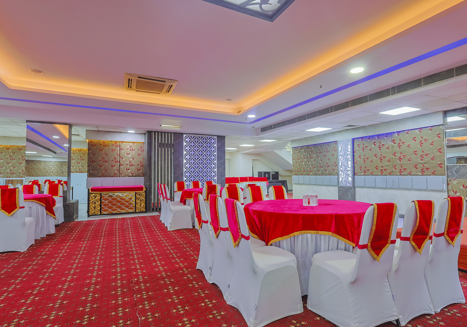  banquet hall set up for an event, featuring tables with white chairs and red tablecloths, arranged on a red patterned carpet.
