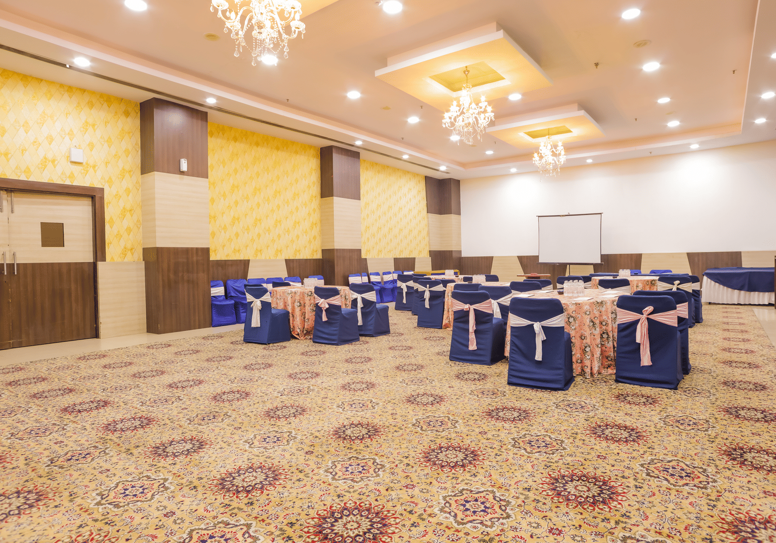 A spacious conference or event room with a patterned carpet, featuring chairs draped in blue covers arranged around tables, and a stage area.