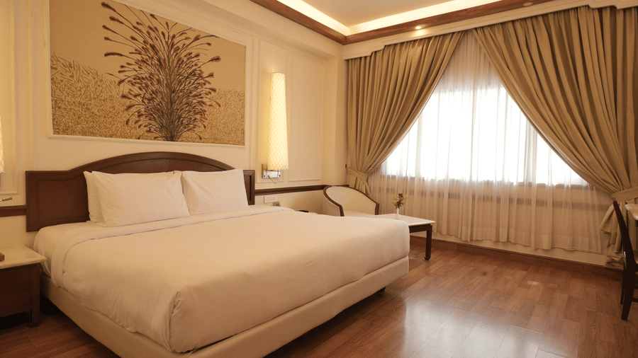 Polished hardwood floors leading towards a bed and large window with draped curtains in the Superior King room at Clarks Amer, Jaipur.