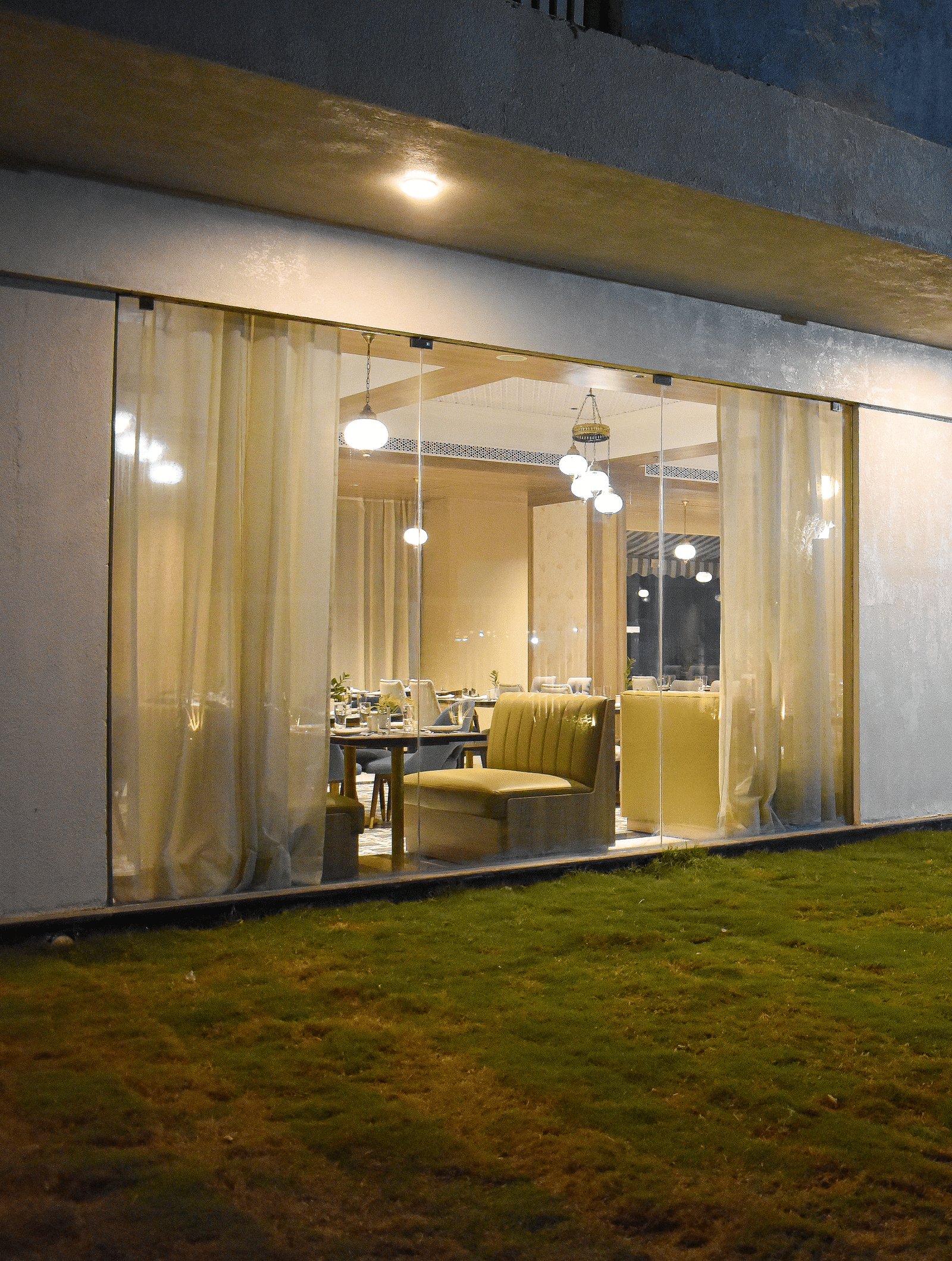 Exterior view of a building with a glass-walled restaurant, sheer curtains, hanging lights, and a grassy area in front - Urban Suites by BluSalzz, Jaipur