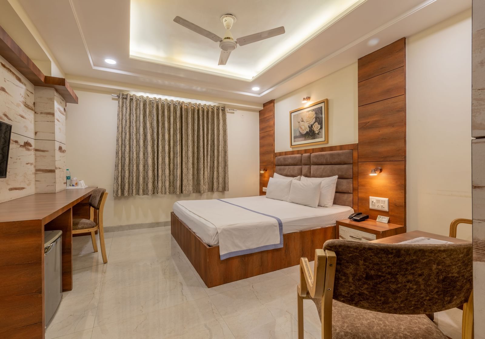 A room featuring a bed with white and blue linen, a wooden desk and chair, and a wall art at Hotel Highway King, Jaipur.