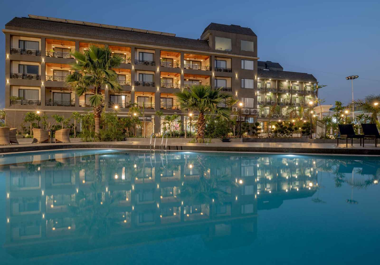  A multi-storey hotel building with colourful window accents overlooks a large, inviting swimming pool with lounge chairs at dusk.