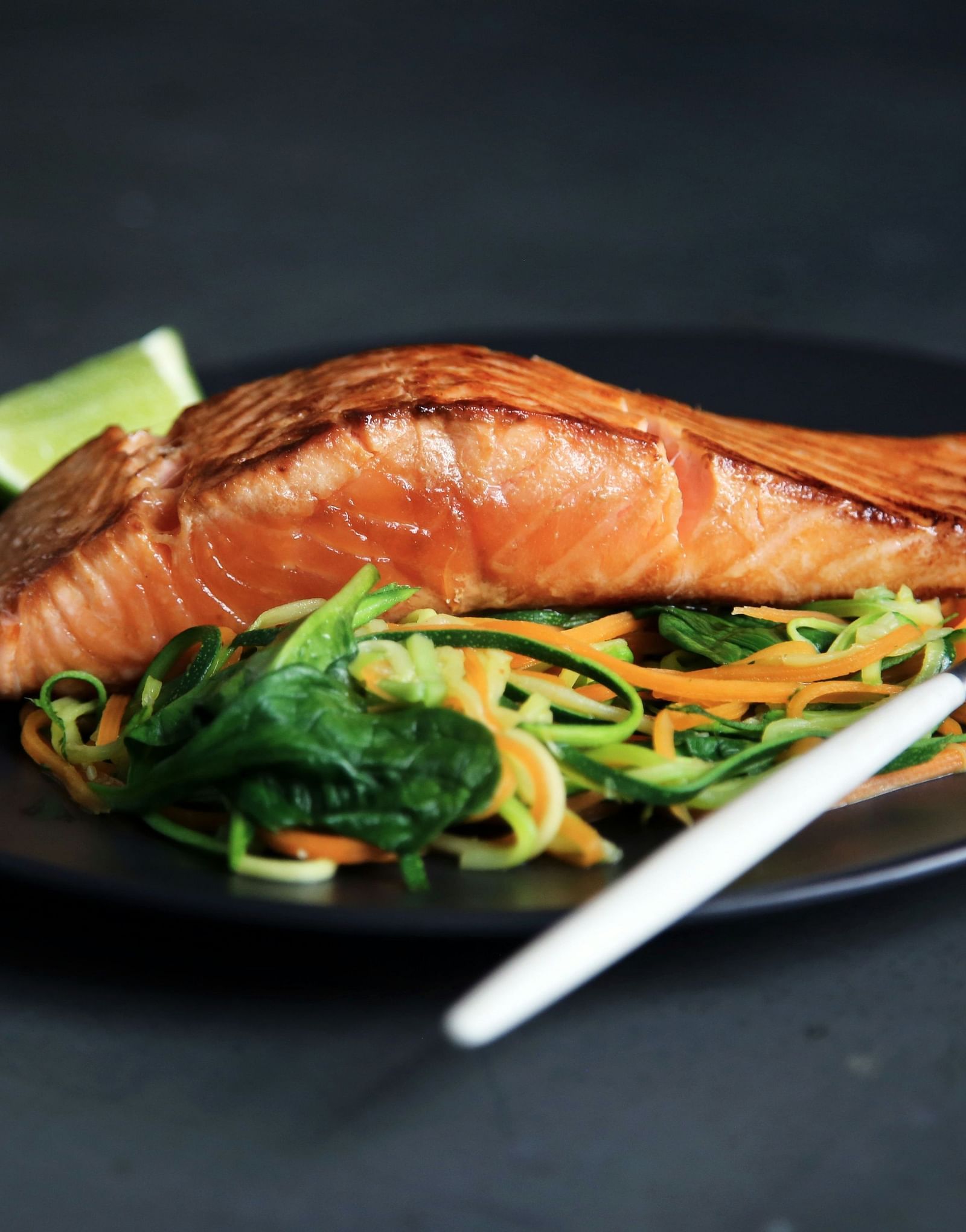 A plated dish of grilled salmon served on a bed of sautéed greens and noodles, with a lime wedge on the side.