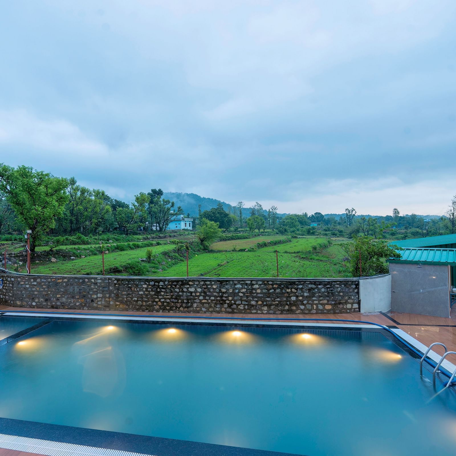 view of the swimming pool at Corbett Four Seasons, Ramnagar, with lush fields and mountains behind it