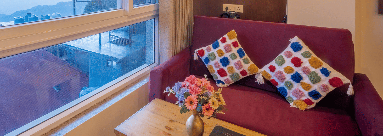 A hotel room with a large window view of a sunset or sunrise over hills, featuring a purple sofa with colourful pillows | Perfectstayz Value Valley Vista
