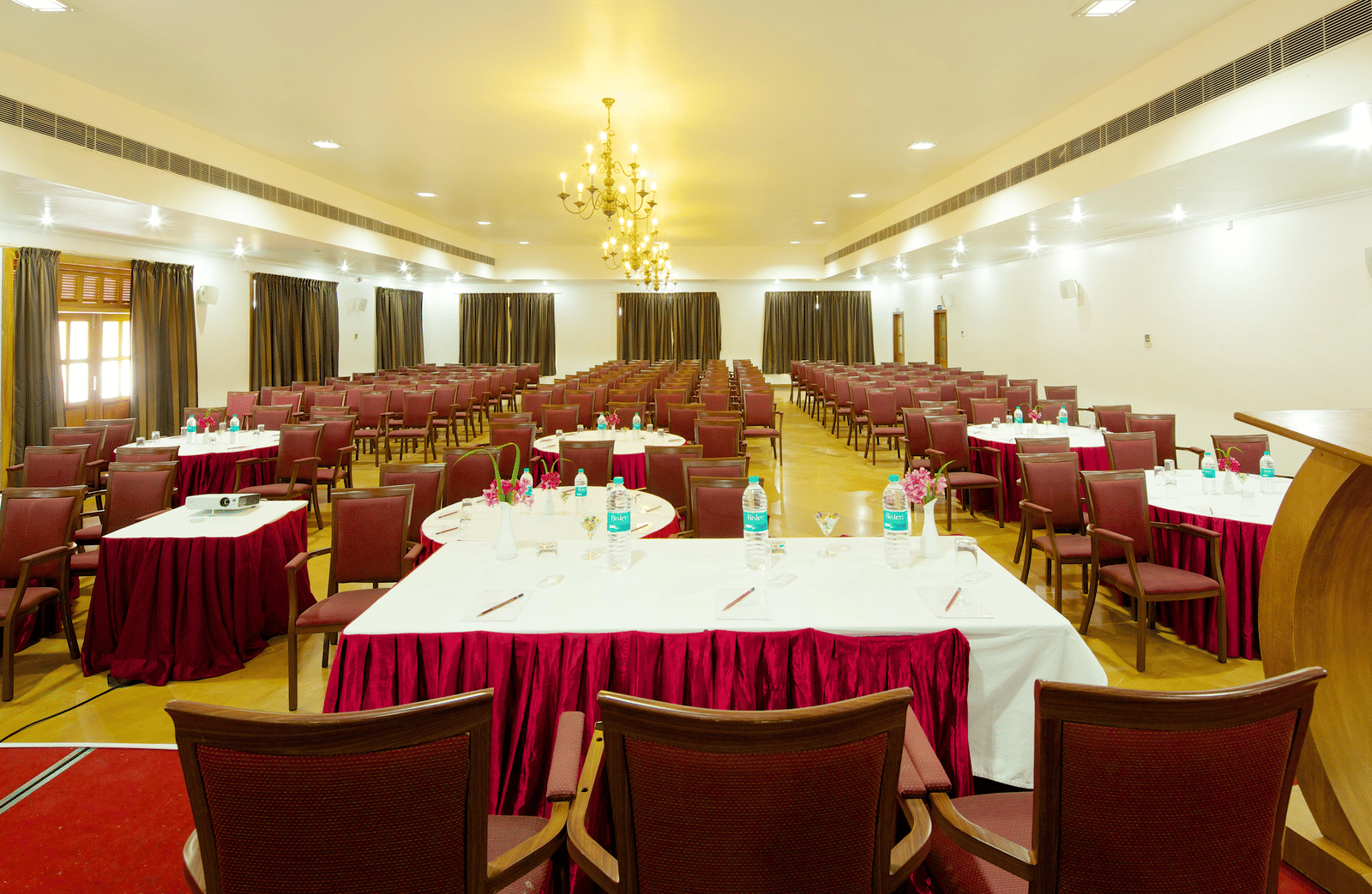 A large meeting room arranged with multiple tables covered in white tablecloths with red valances and surrounded by chairs | Le Pondy