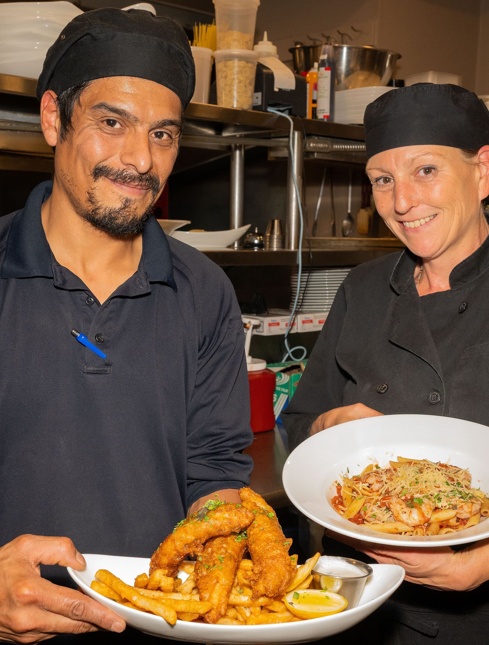 Two chefs smiling while holding plated dishes in the kitchen at The Groveland Hotel.