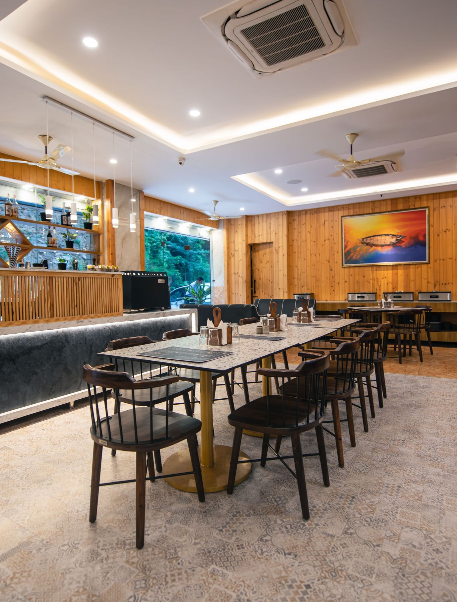A restaurant at The Tattva Devaprayag with a long wooden table with black chairs and a bar.