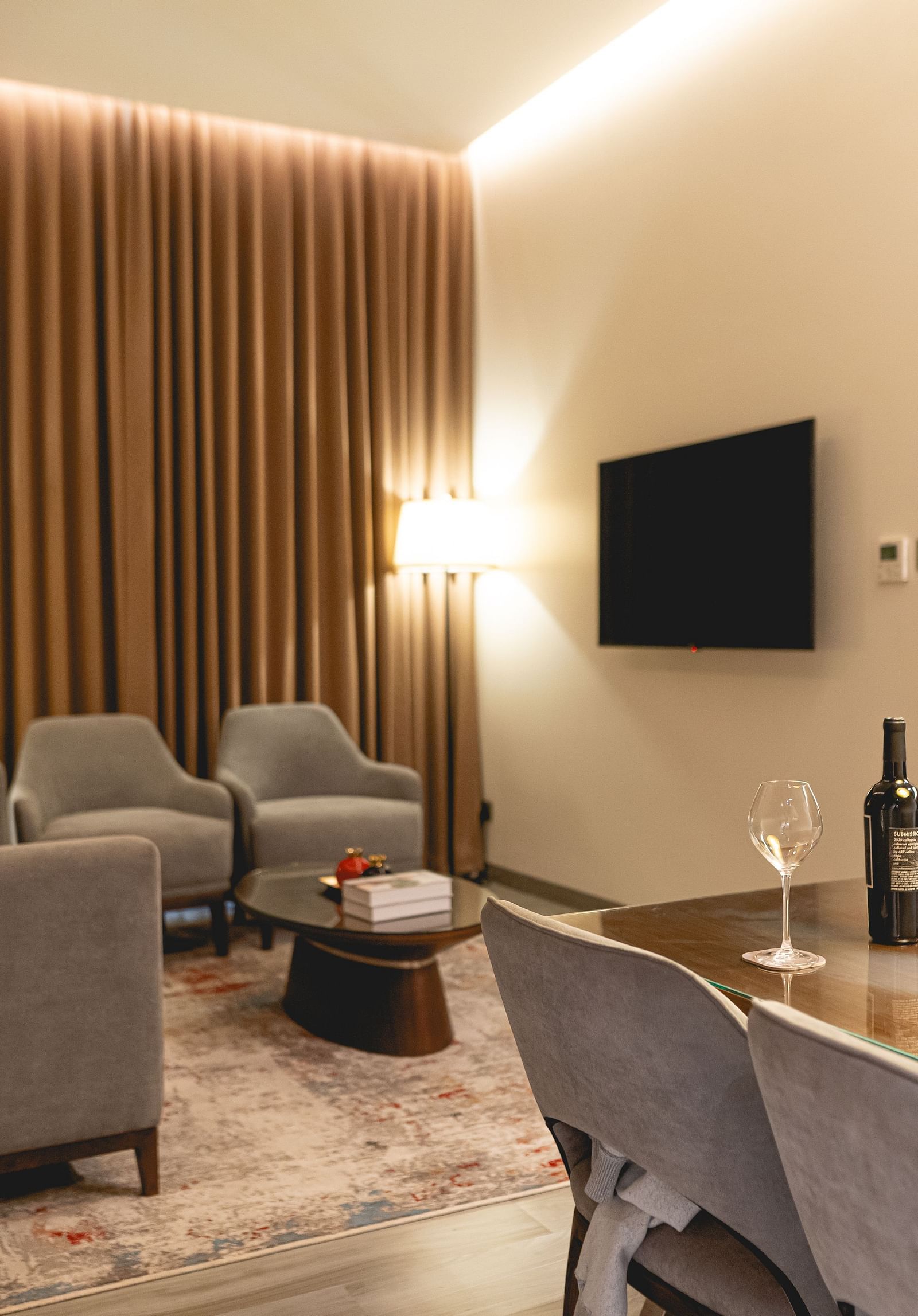 Modern living room at KertelSuites with neutral tones, featuring a sofa, armchairs, TV on the wall, floor and table lamps, and a dining table with chairs; warm lighting and vertical wooden slats on one wall