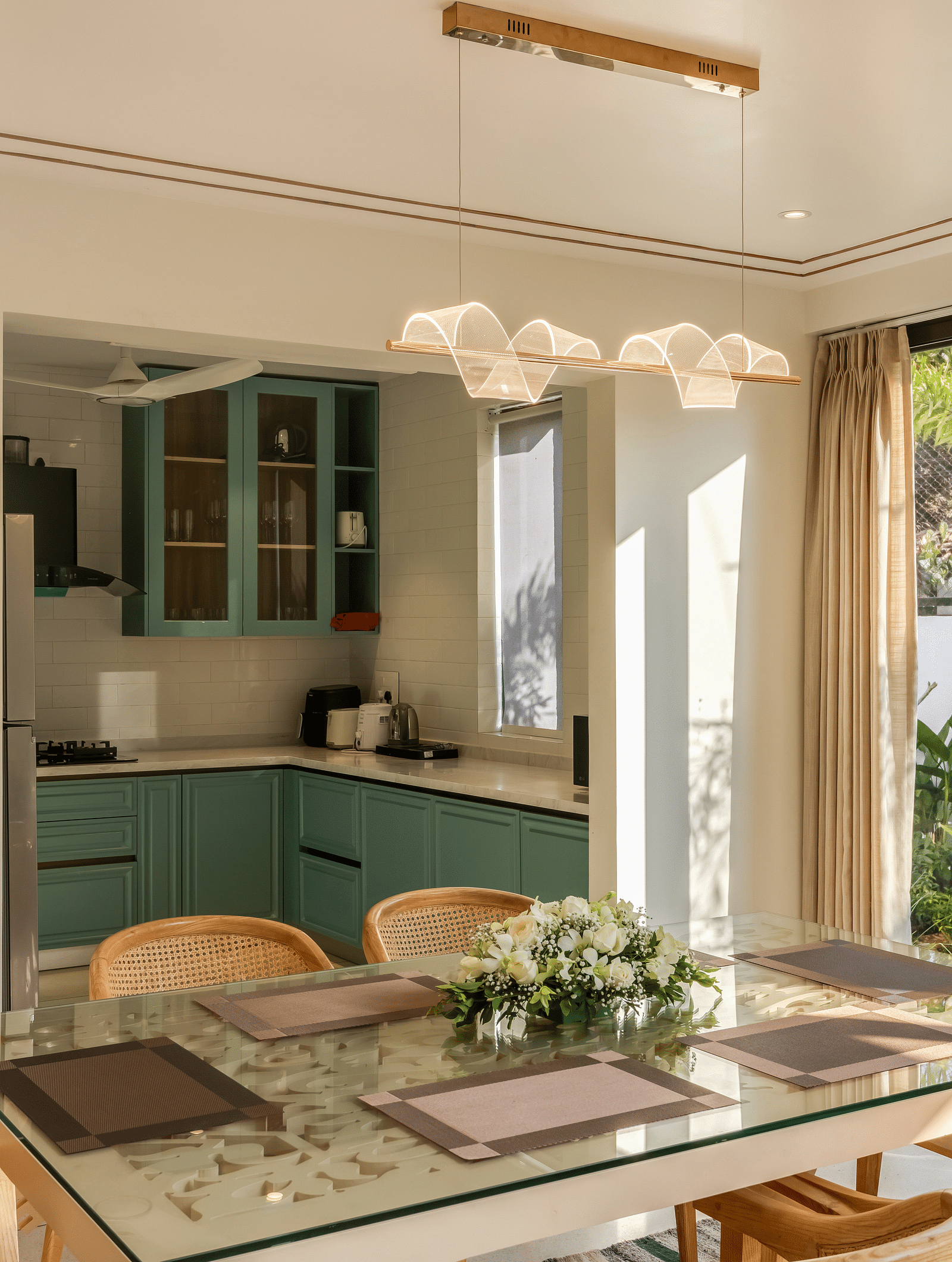 A dining area with a modern wooden table and chairs, adjacent to a bright kitchen with a large window offering a lush outdoor view.