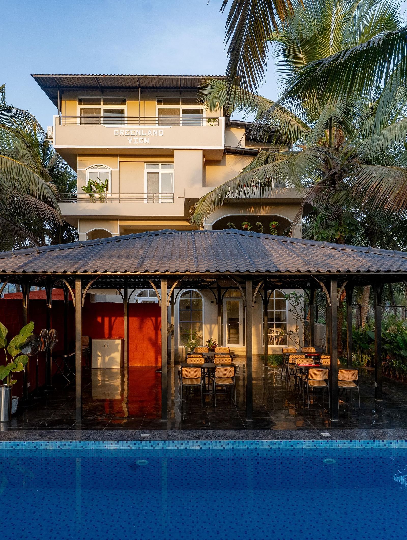 A view across a swimming pool towards a covered outdoor seating area with a thatched roof. Palm trees sway gently in the background at Kssemya.