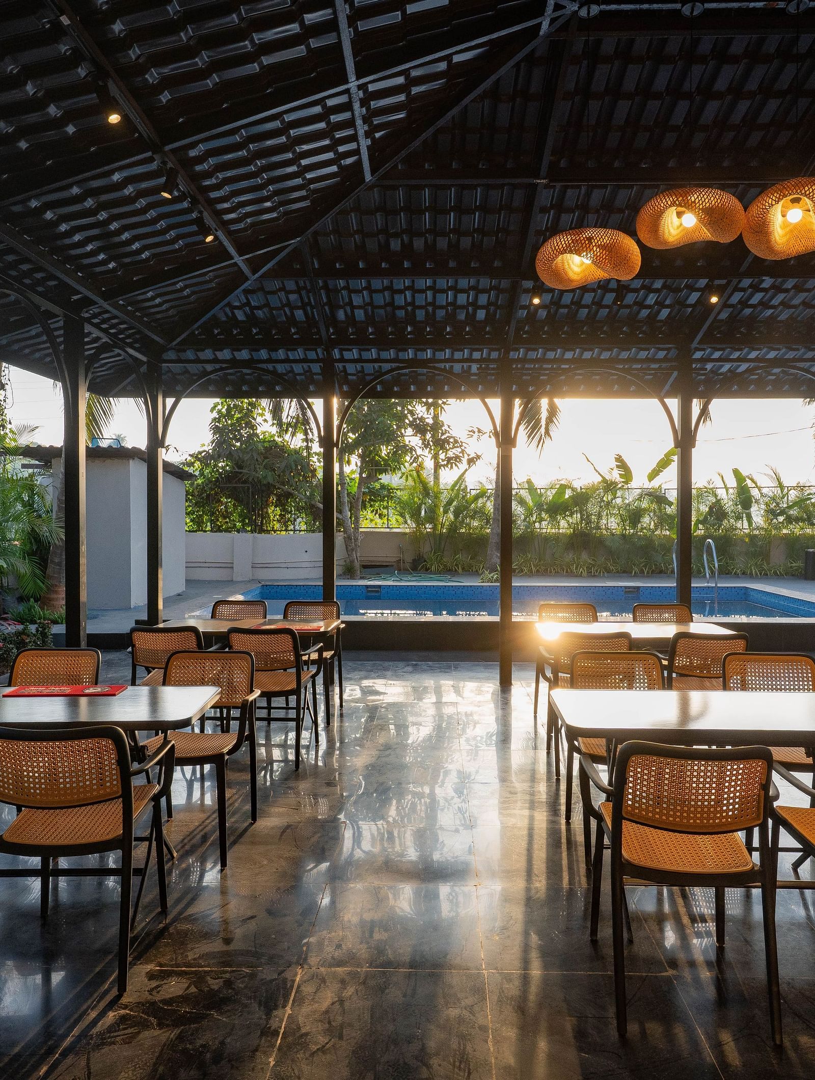 An outdoor dining area under a dark, peaked roof, open to the surrounding scenery. Several wooden tables and chairs are neatly arranged, with a glimpse of a swimming pool visible in the background under the bright sky at Kssemya