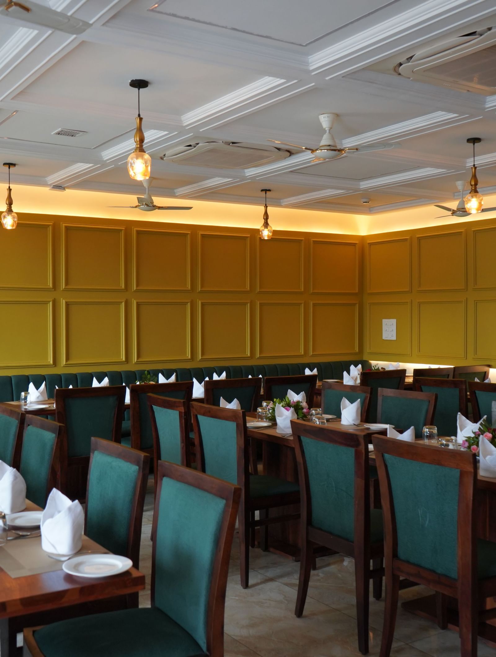 A wide shot of a dining hall with multiple tables set for a meal. The chairs are dark green, and the walls are yellow. A TV is visible on the far wall, showing a soccer game.