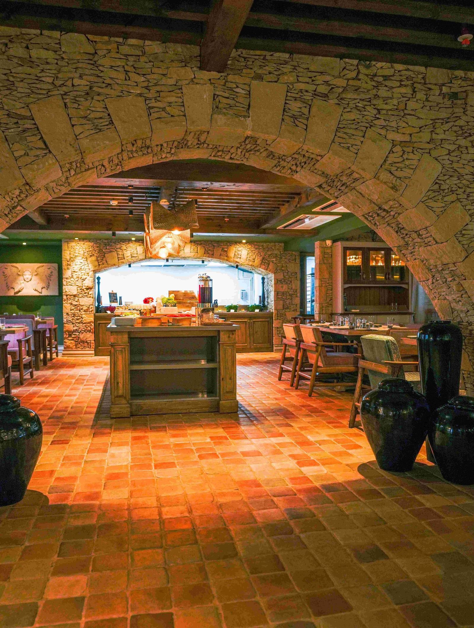 A view into Mila Restaurant at The Evren, Vagator, taken through a rustic stone archway. The restaurant has a tiled floor, a central buffet area, and various dining tables, creating a warm and inviting atmosphere.