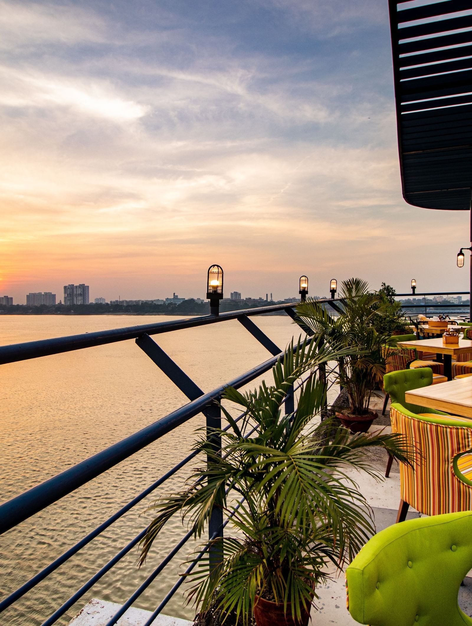 A deck on a river overlooking the city at sunset with tables surrounded by green and yellow cushions - Polo Floatel Kolkata