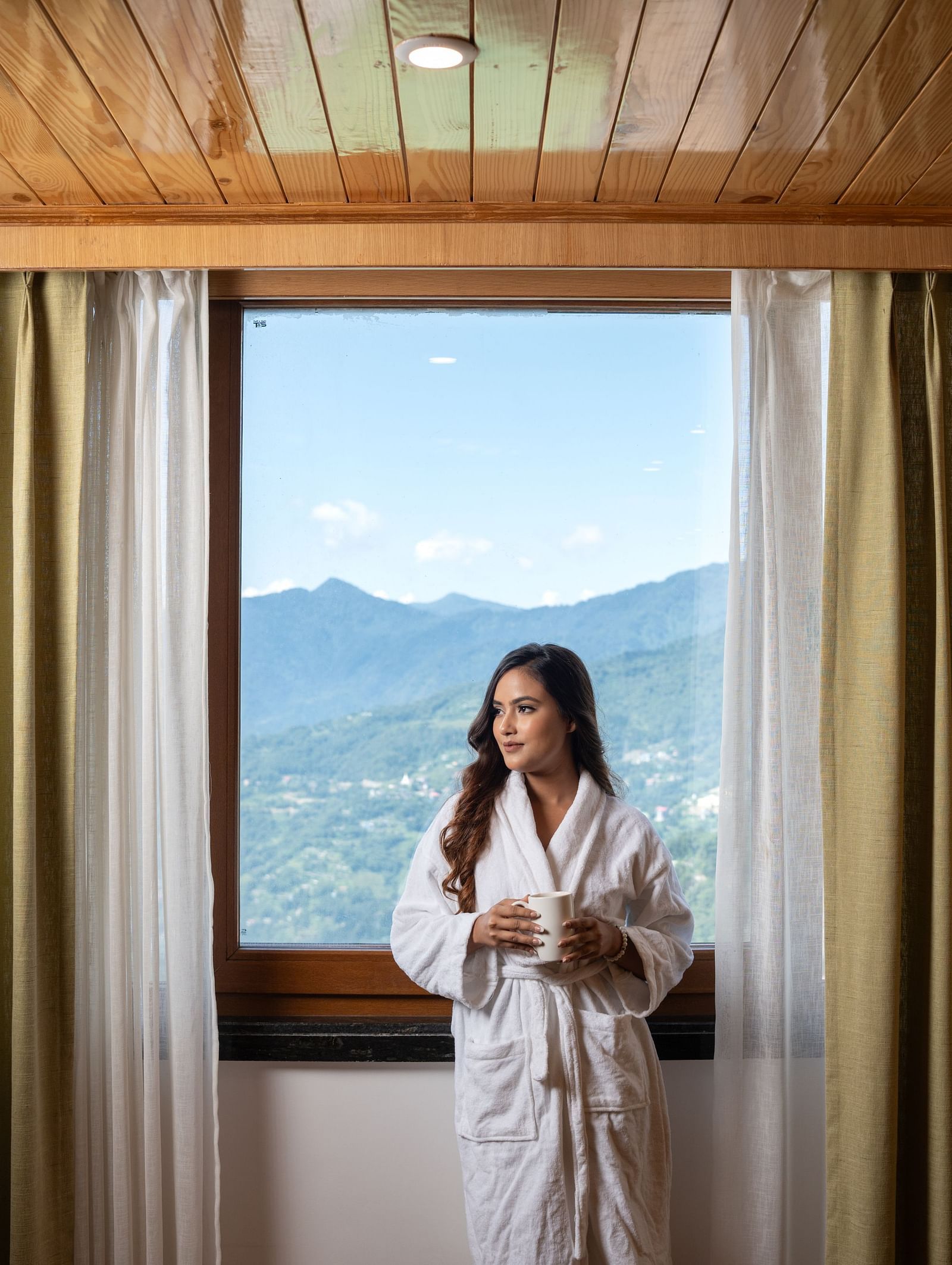 A lady stands by the window, curtains drawn aside, with mountains in the background