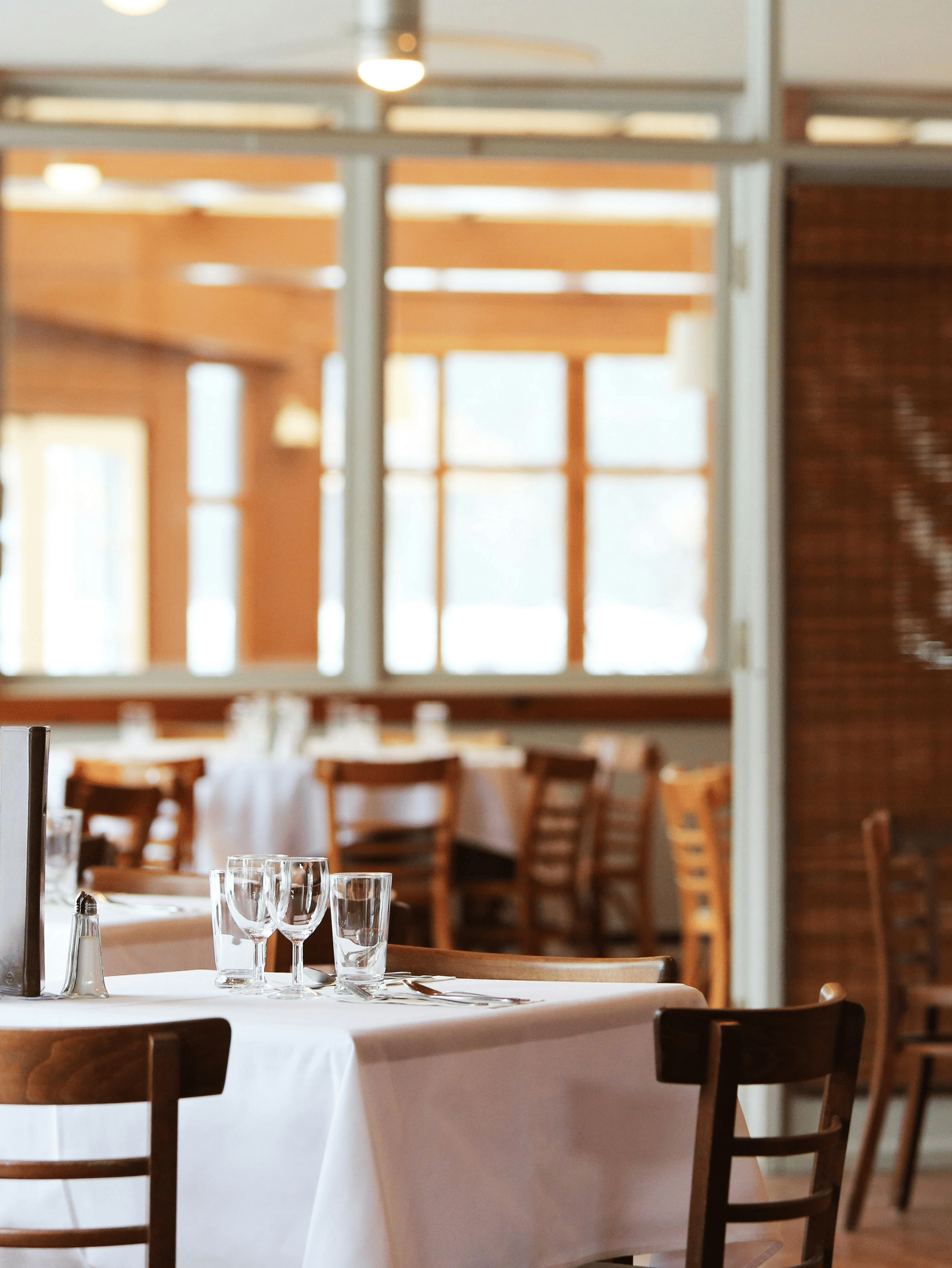 The bright interior of a restaurant with wooden tables and chairs, crisp white tablecloths, and bamboo window blinds.