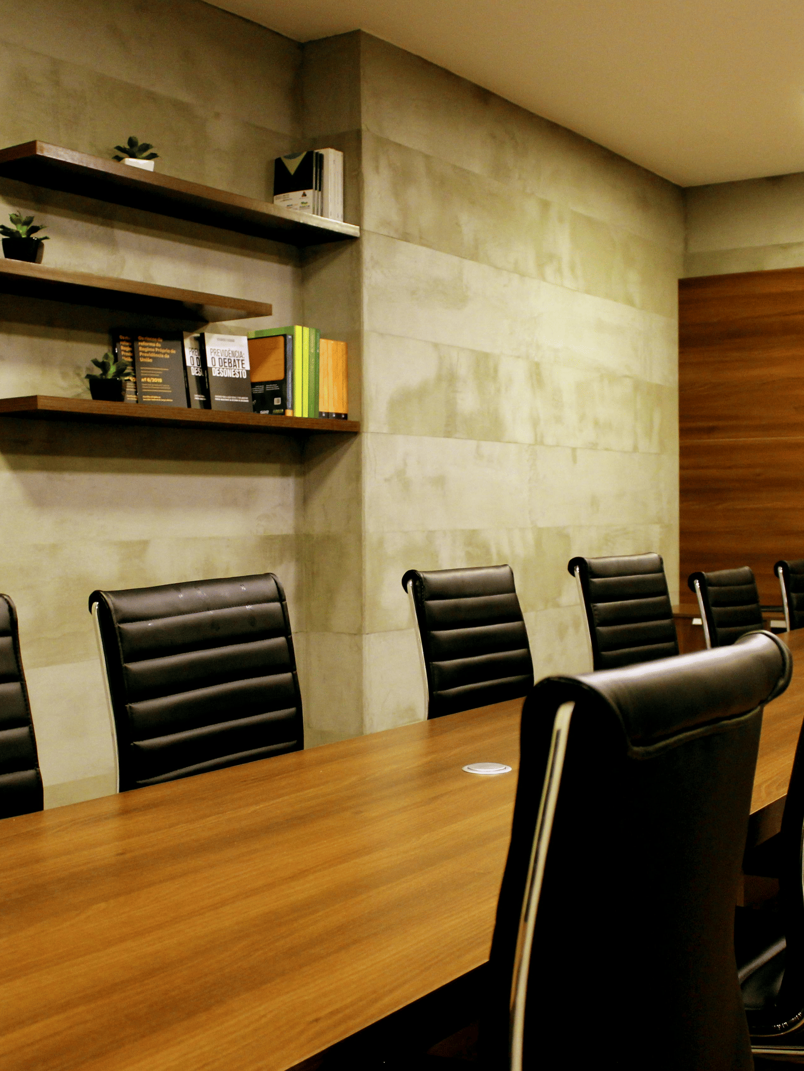 A corporate board room with a large wooden table, several black leather executive chairs, and floating shelves on a stone wall.