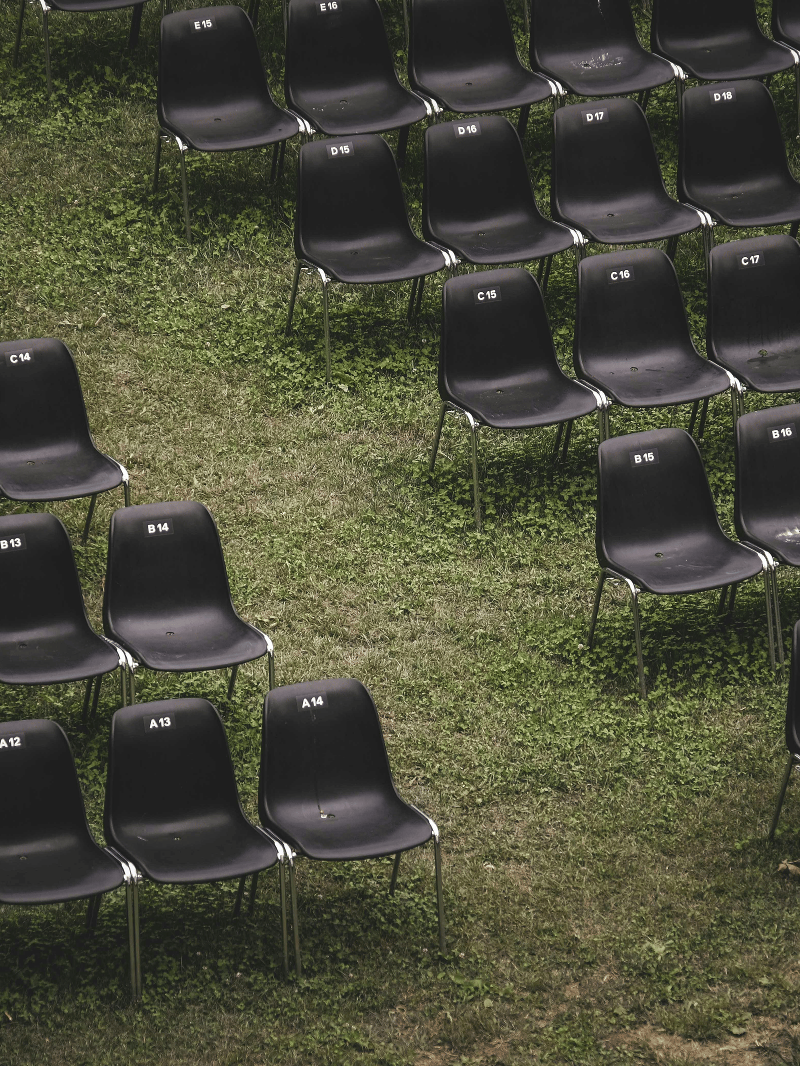 Rows of empty, dark plastic chairs set up in an open-air venue on a slightly overgrown green grass lawn.