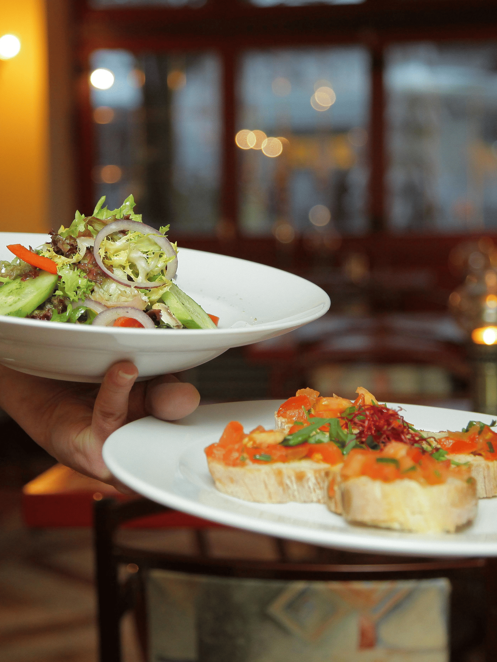 A waiter in a white shirt and black waistcoat is serving two plates of food, one with a side salad and one with bruschetta.