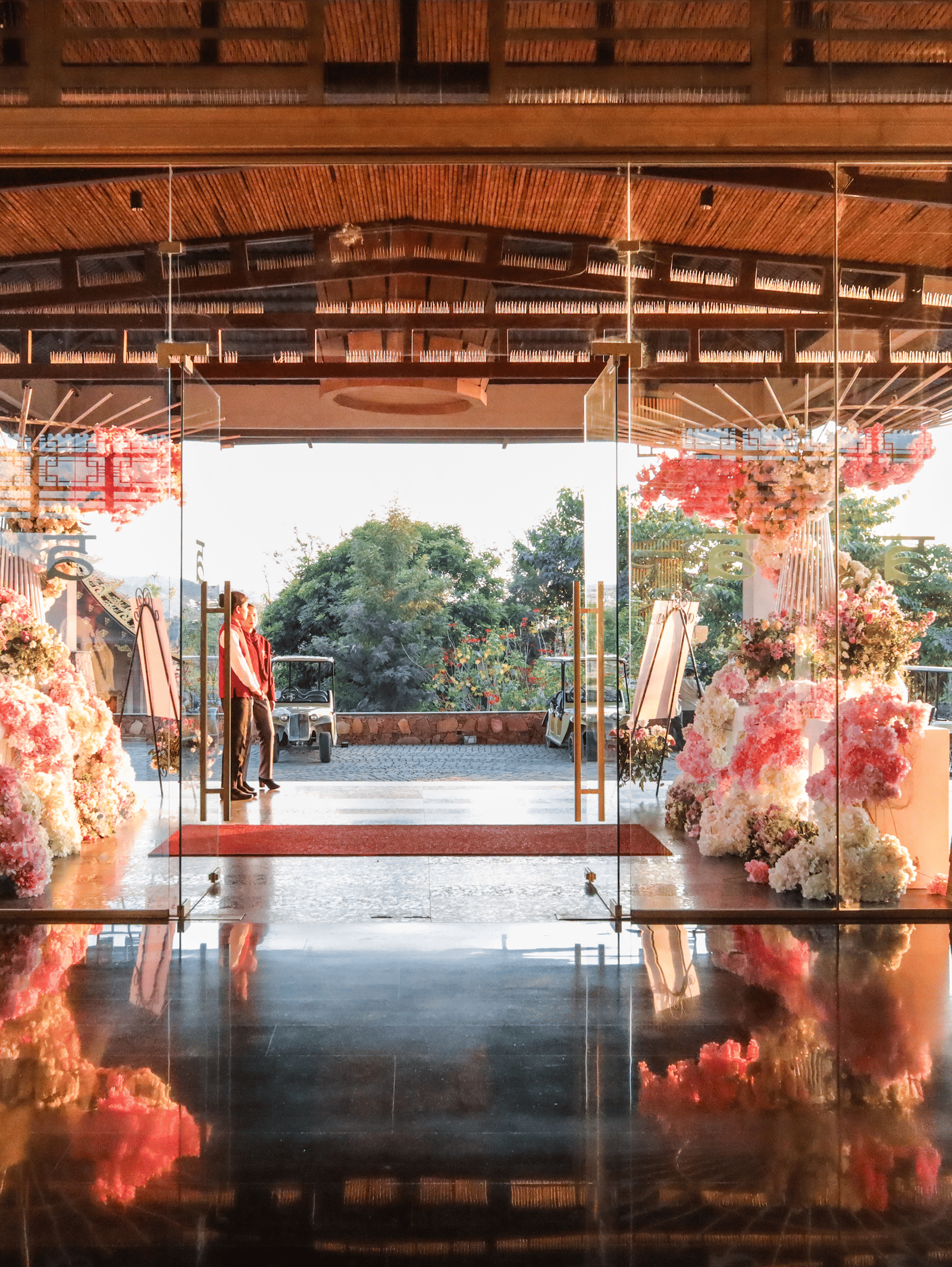 Attendants stand inside the glass entrance of Ananta Spa & Resort, Jaipur, which is decorated with large pink and white floral arrangements.