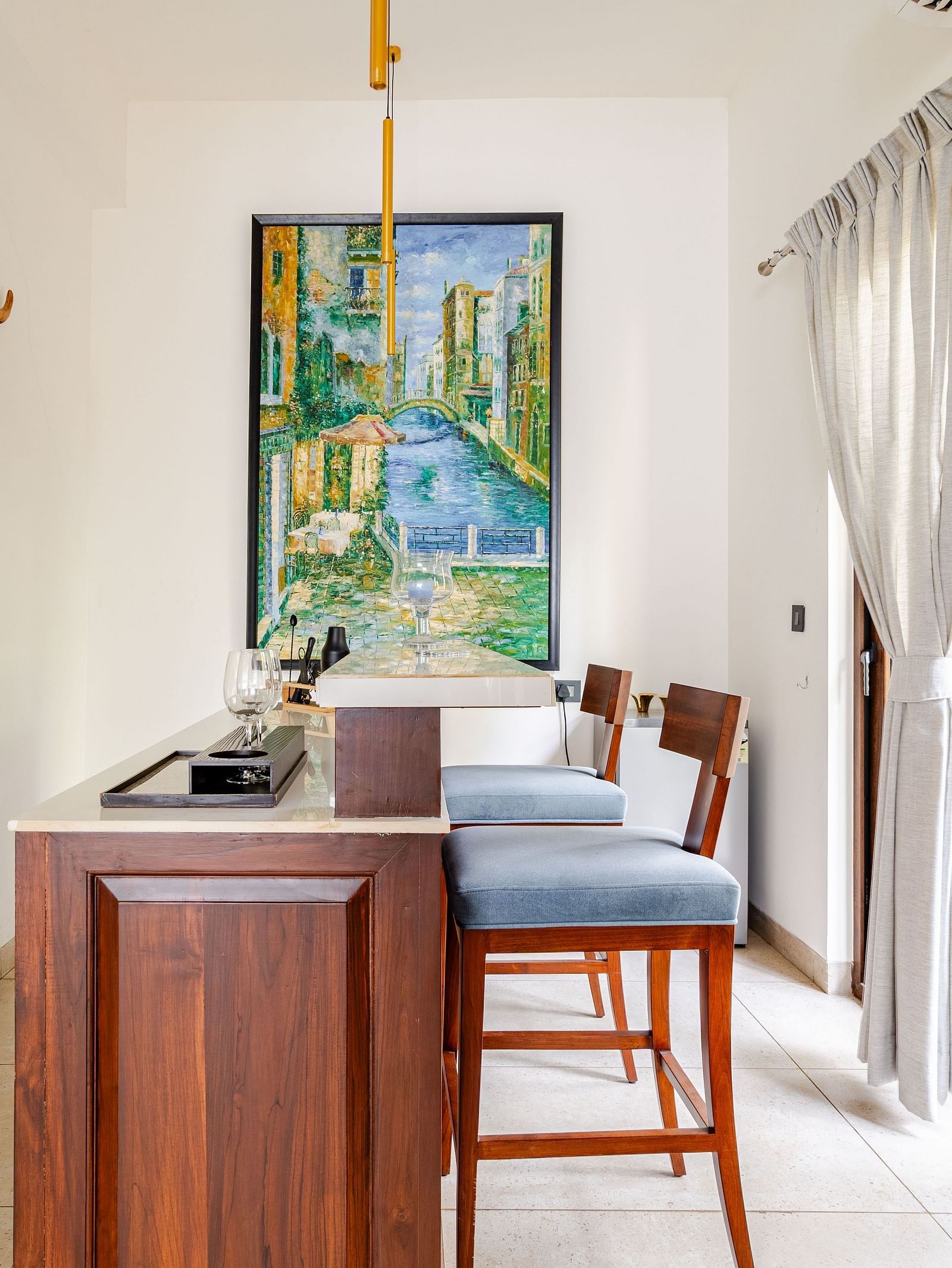 A bar area featuring a compact wooden counter with two high chairs, a colourful canal-themed painting, decorative bull skull wall art, and sheer curtains allowing natural light through large glass doors with a view of green foliage outside - Lindsay Manor
