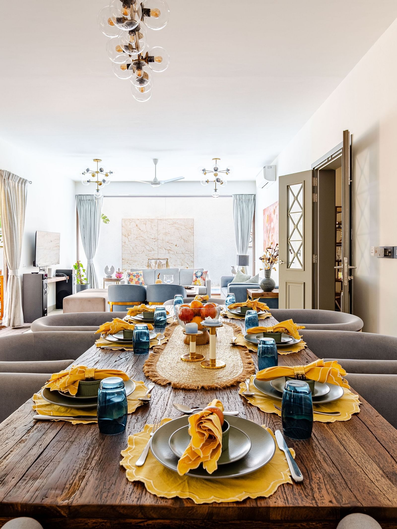 Elegant open-plan dining and living area featuring a rustic wooden table set with yellow napkins and blue glasses, surrounded by plush grey chairs, with natural light streaming in through sheer curtains and glass doors leading to an outdoor seating area - 