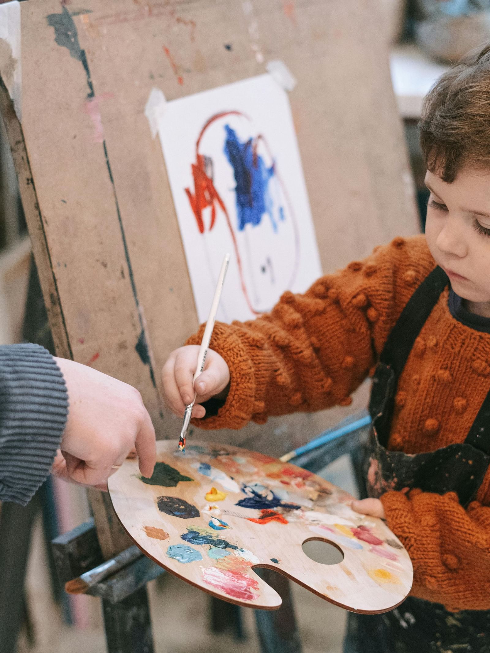 A young child intently paints on an easel guided by an adult holding a palette.