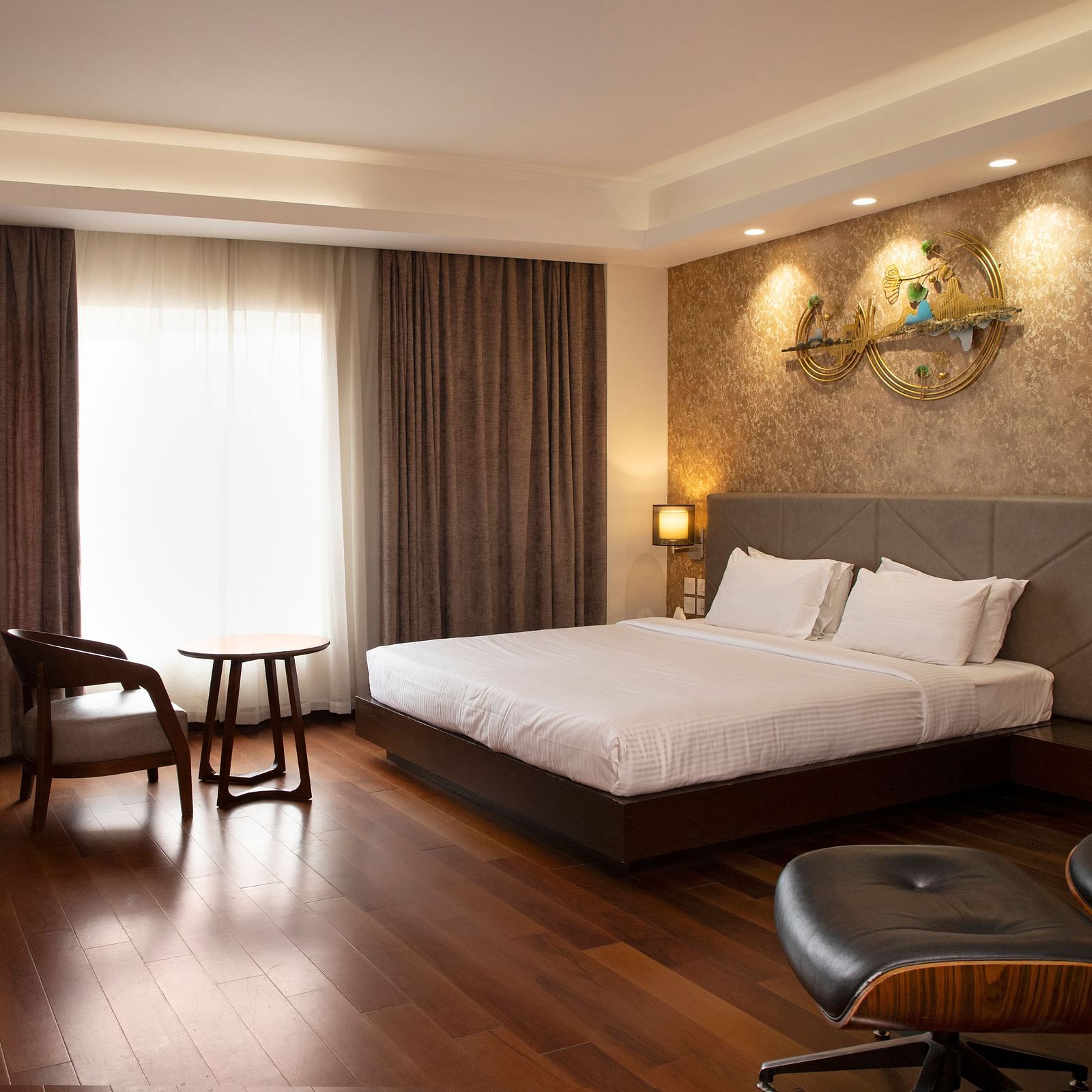 A spacious room featuring a large bed, a cosy seating area, elegant curtains, soft ambient lighting, and expansive windows with curtains drawn to the side at The Hamuse Hotel, Kodaikanal.