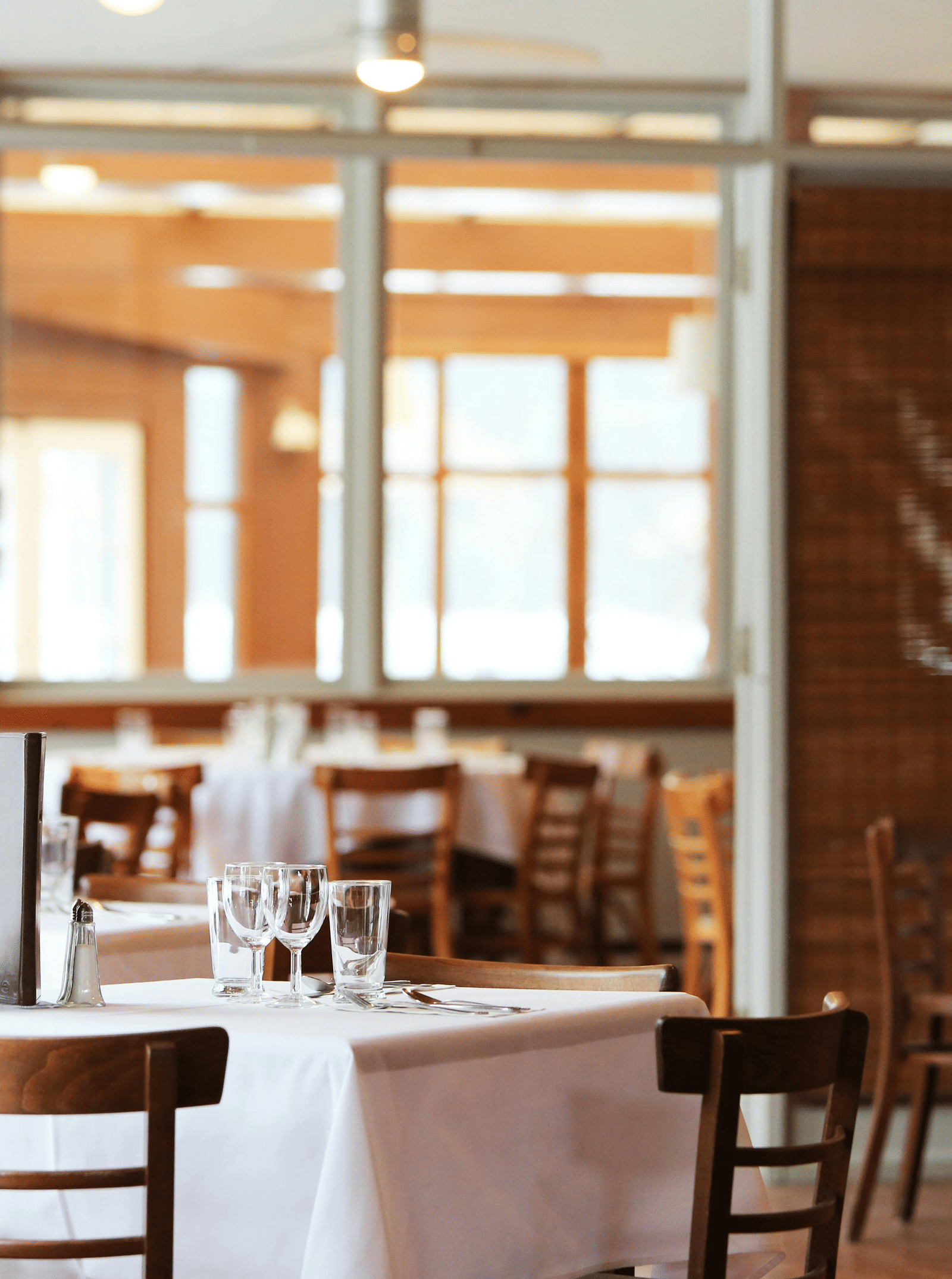 The bright interior of a restaurant with wooden tables and chairs, crisp white tablecloths, and bamboo window blinds.