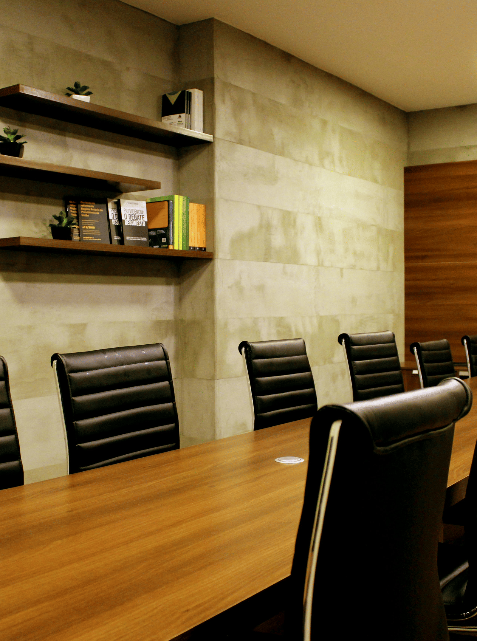 A corporate board room with a large wooden table, several black leather executive chairs, and floating shelves on a stone wall.