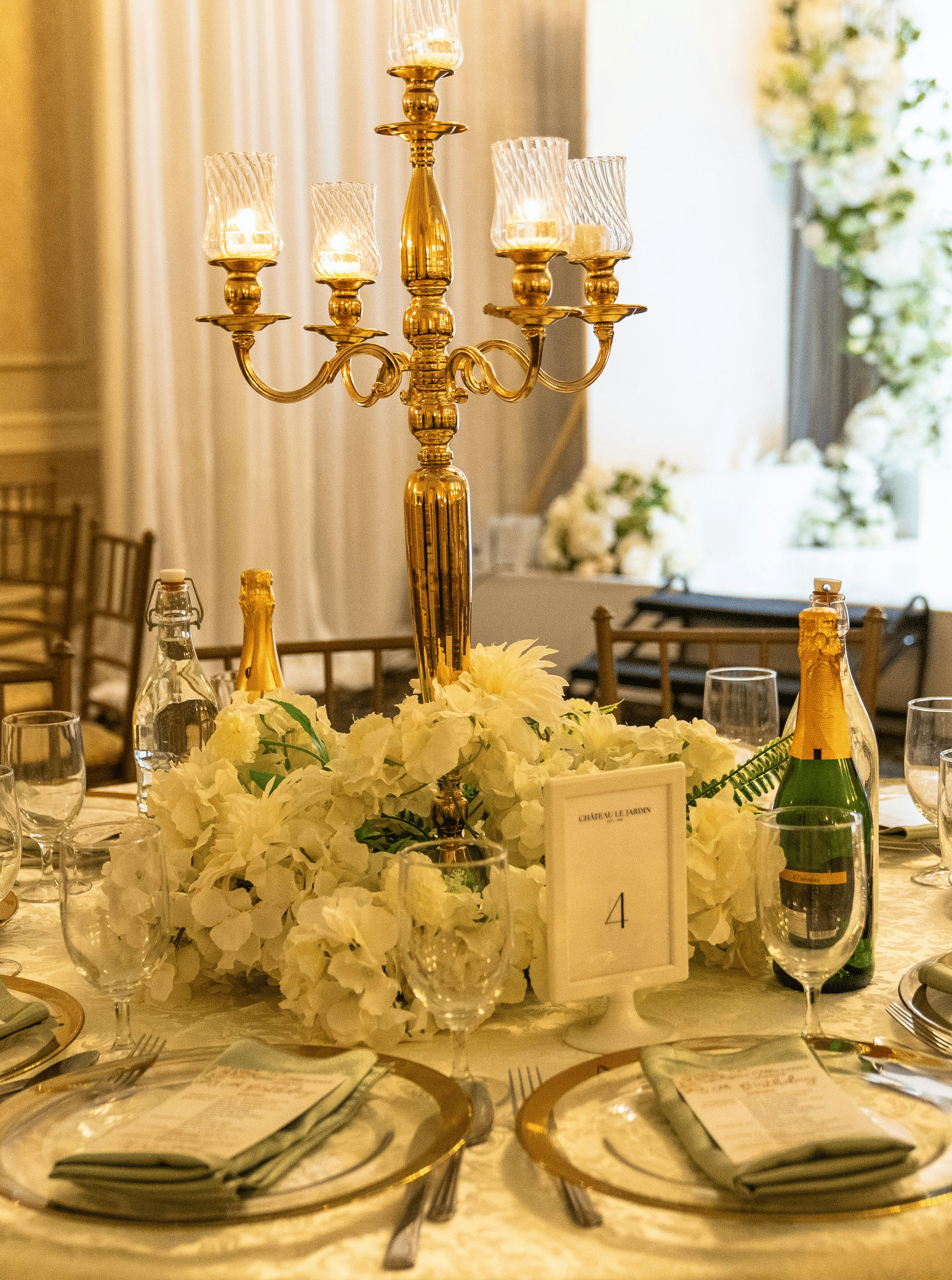 A formal dining table set for a wedding or event, featuring a large hydrangea centrepiece and a gold candelabra.